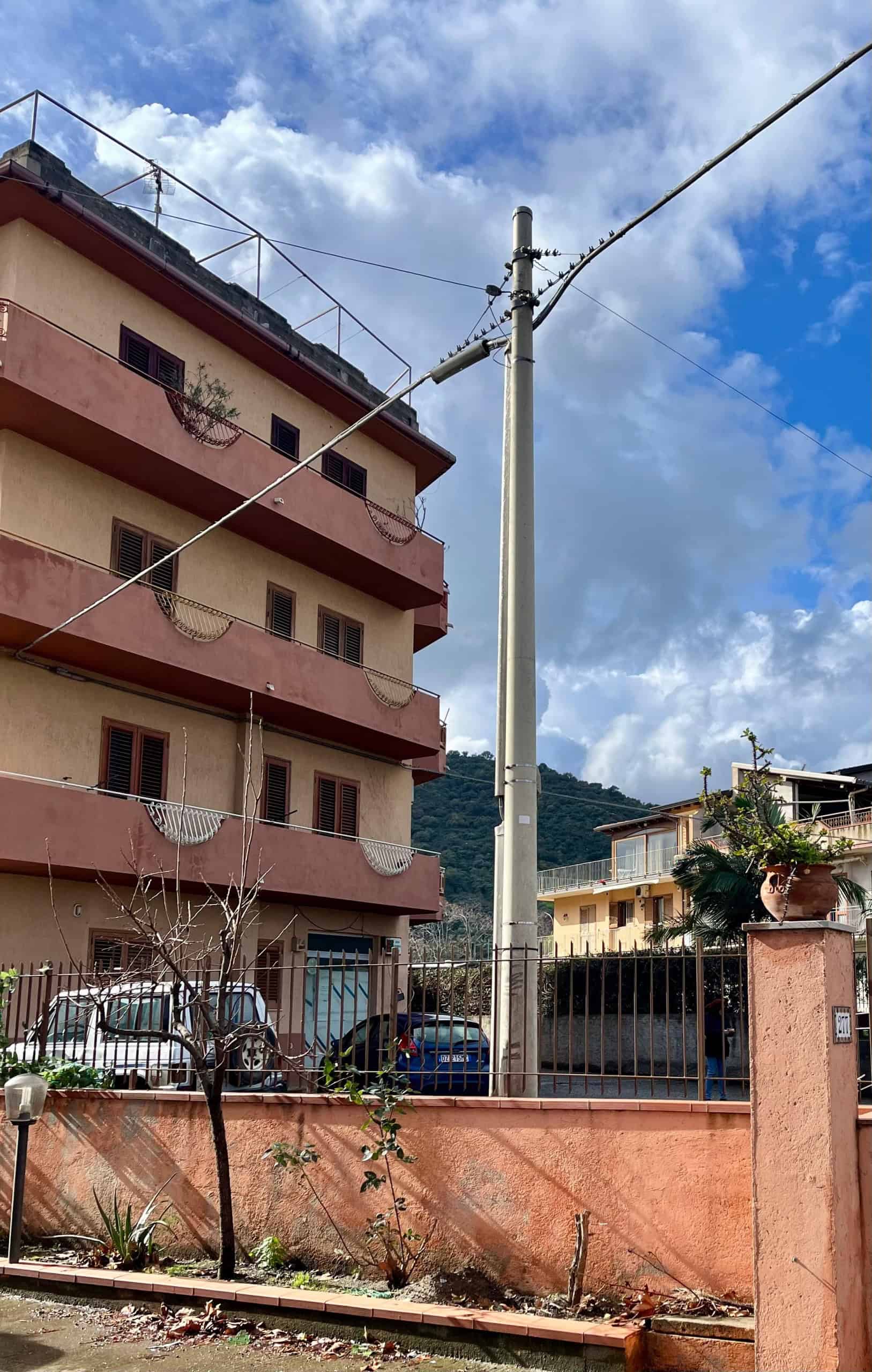Palo della luce con cavi elettrici in un quartiere residenziale, con edificio a più piani e montagne sullo sfondo, cielo nuvoloso, in Italia.
