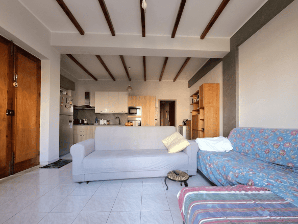 Soggiorno luminoso con divano bianco e divano letto con copertura floreale, in un appartamento in stile rustico moderno, con soffitto a travi di legno e pareti chiare, ambiente accogliente e spazioso.