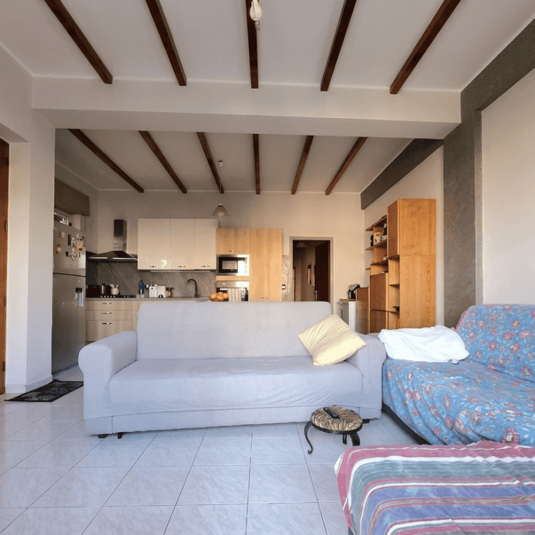 Soggiorno luminoso con divano bianco e divano letto con copertura floreale, in un appartamento in stile rustico moderno, con soffitto a travi di legno e pareti chiare, ambiente accogliente e spazioso.