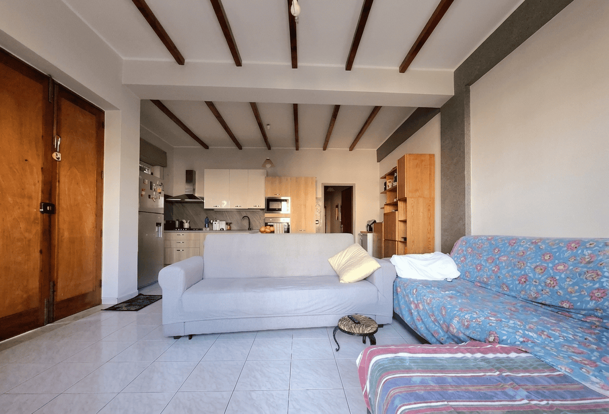 Soggiorno luminoso con divano bianco e divano letto con copertura floreale, in un appartamento in stile rustico moderno, con soffitto a travi di legno e pareti chiare, ambiente accogliente e spazioso.