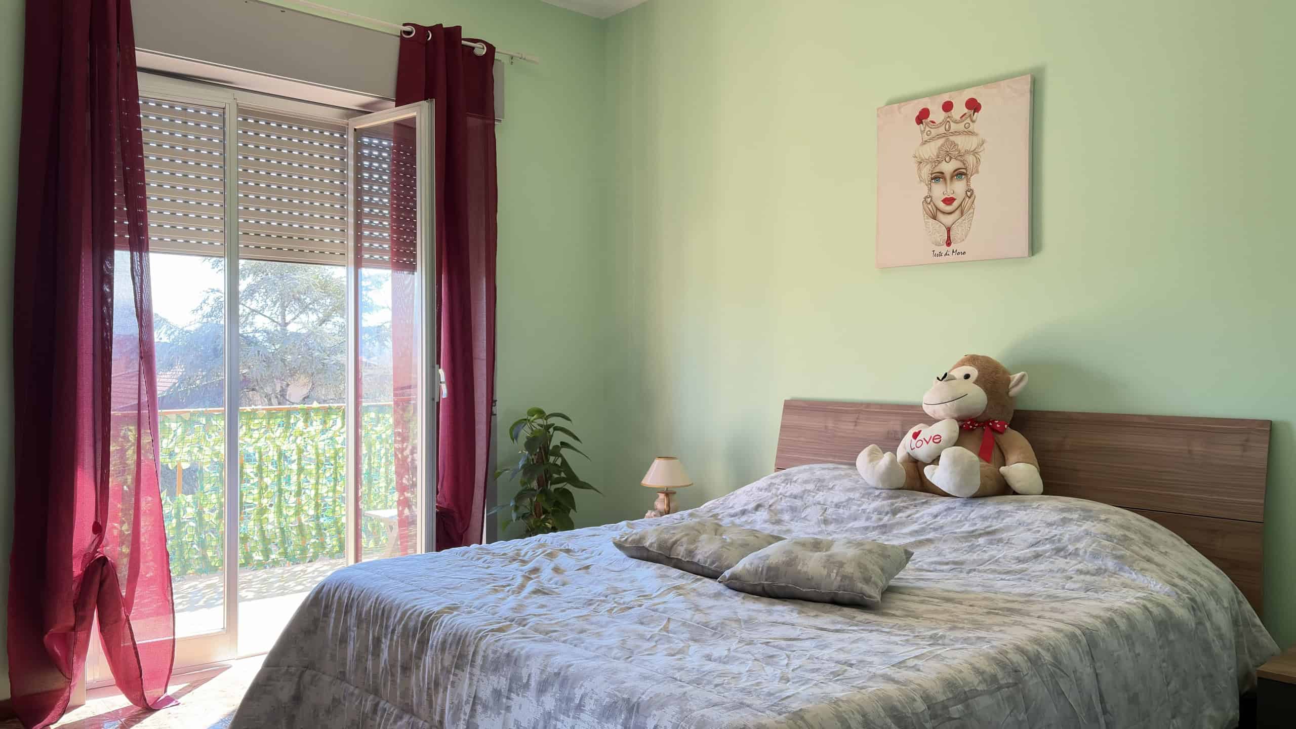 Letto matrimoniale con cuscini e orsacchiotto in una camera luminosa con pareti verde pastello, balcone con vista su giardino, decorazioni moderne e atmosfera accogliente.