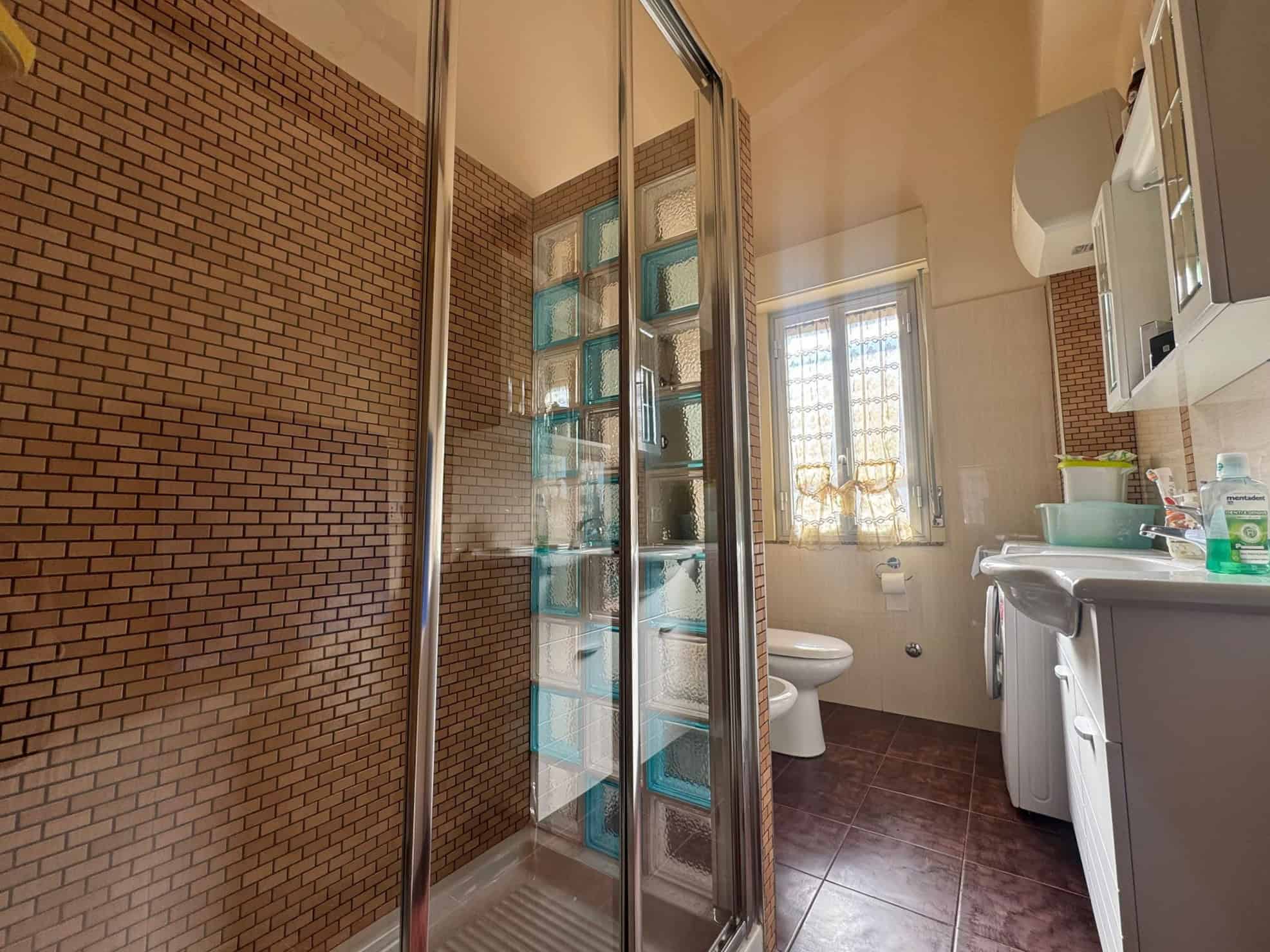 Doccia a filo con pareti in vetro e piastrelle marroni, bagno luminoso con finestra, lavabo e servizi igienici, arredamento funzionale e moderno, ideale per appartamenti e case in vendita o affitto.