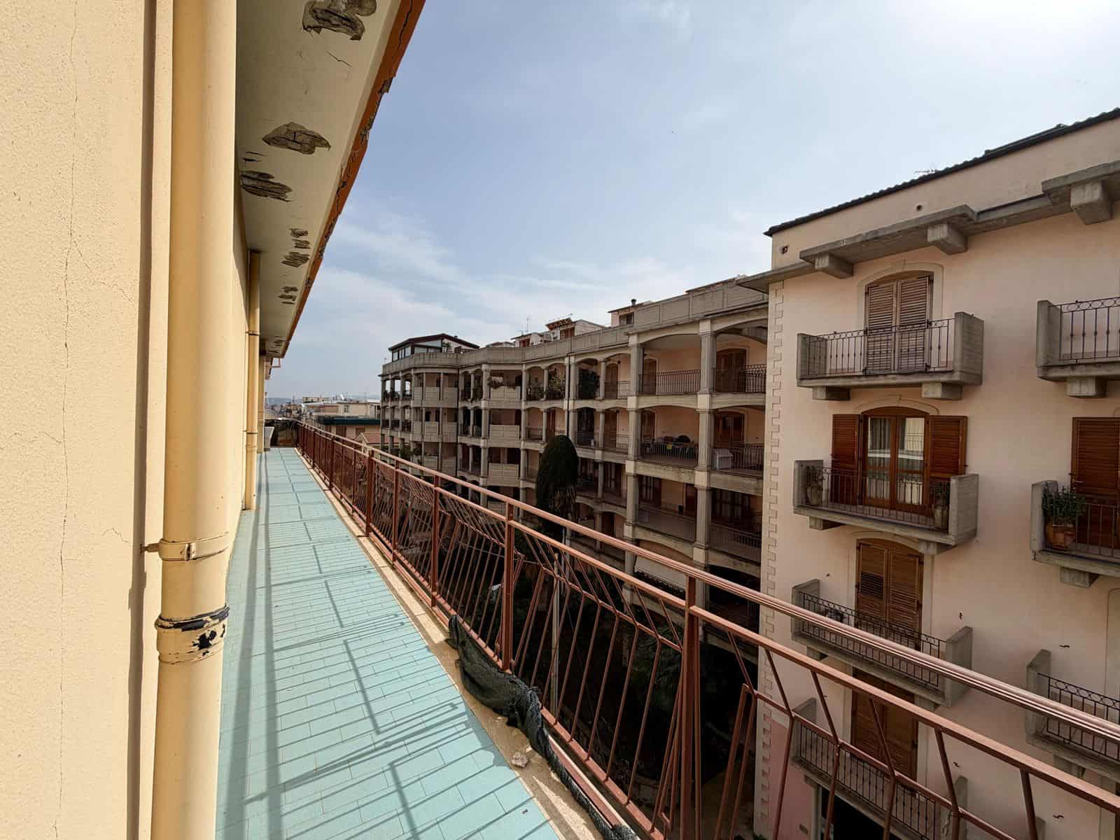 Balcone di un appartamento con vista su edifici residenziali, in un complesso urbano, sotto un cielo sereno, con ringhiera in metallo e pavimentazione in piastrelle azzurre, in un contesto di città.