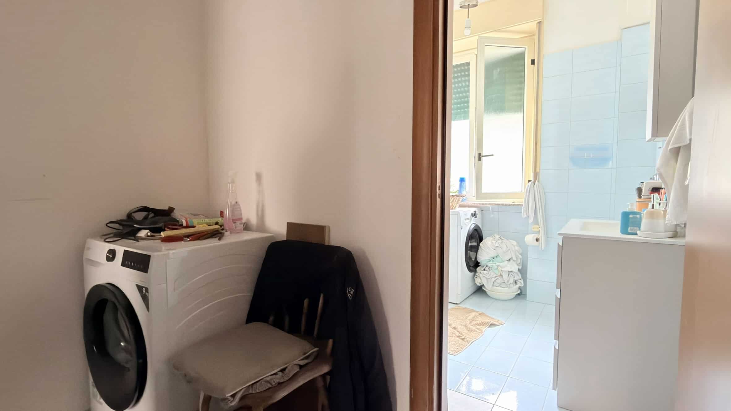 Lavatrice moderna in un ambiente domestico con accessori e detersivi, vicino a una sedia con vestiti piegati, in una casa luminosa e ben organizzata.