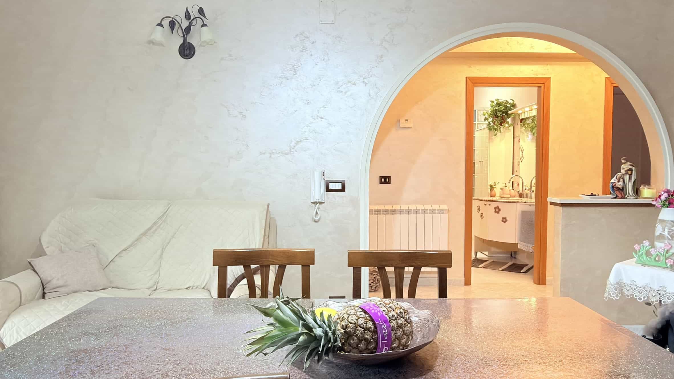 Ampia sala soggiorno con divano bianco, tavolo in legno e arredo elegante, vista sull'area cucina attraverso un arco, ambiente luminoso e accogliente, ideale per vivere momenti di relax e convivialità.