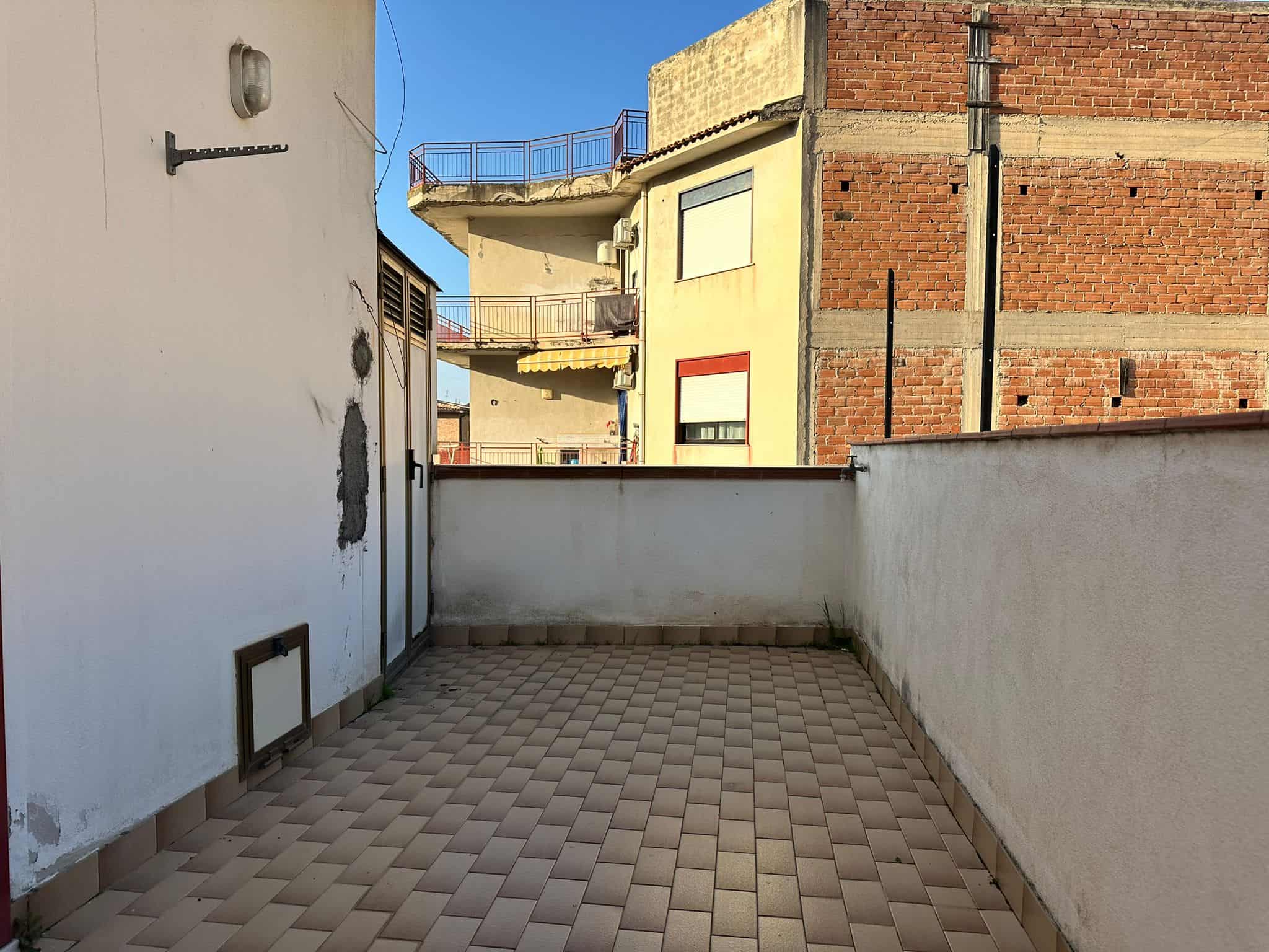 Ampio terrazzo all'aperto con vista su edifici residenziali, pavimentato con piastrelle beige, ideale per relax e socializzazione in zona urbana.