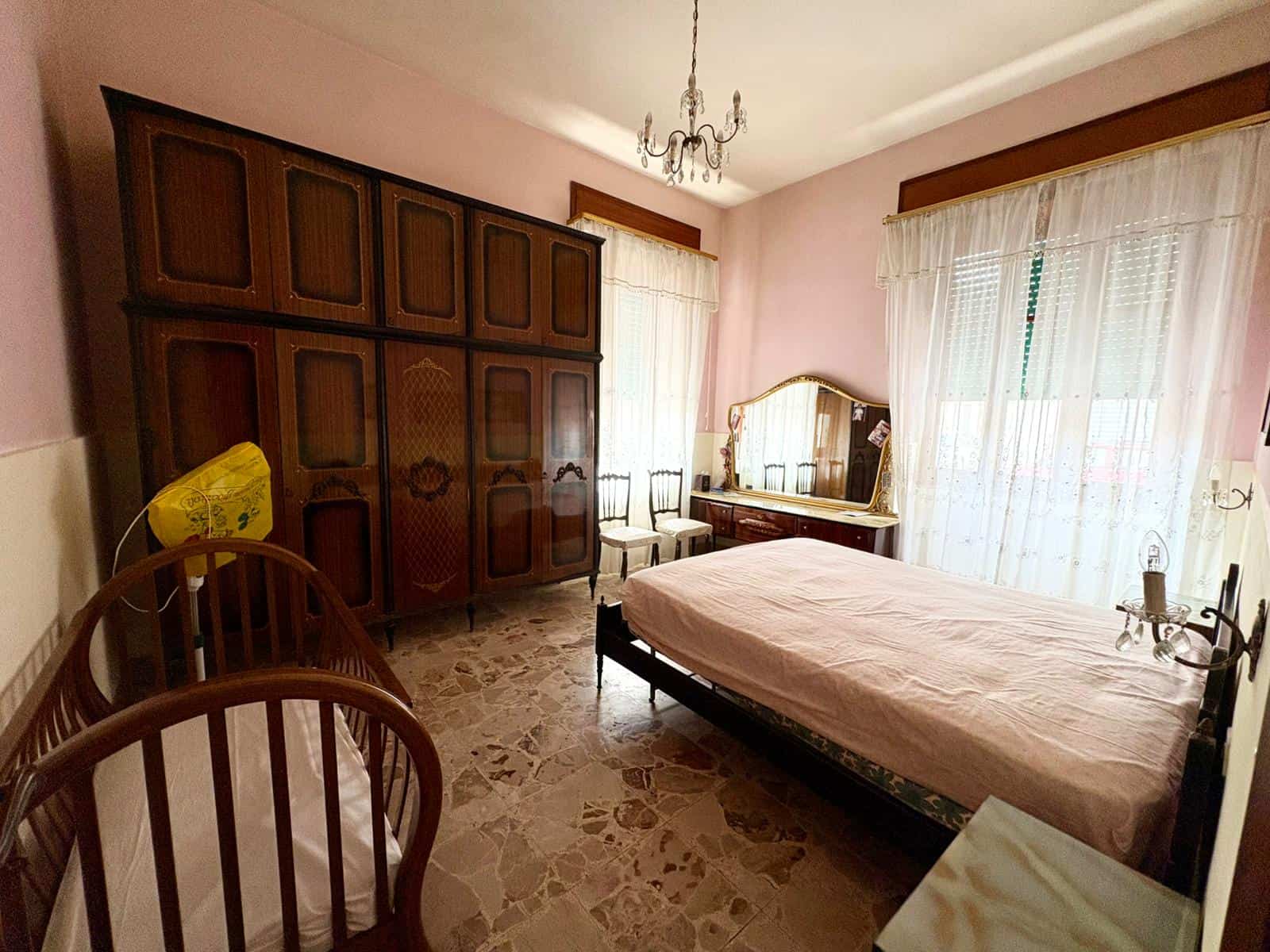 Letto matrimoniale in camera da letto con armadio antico, comò e specchio, illuminata da luce naturale attraverso tende leggere, in un ambiente accogliente e ben arredato.