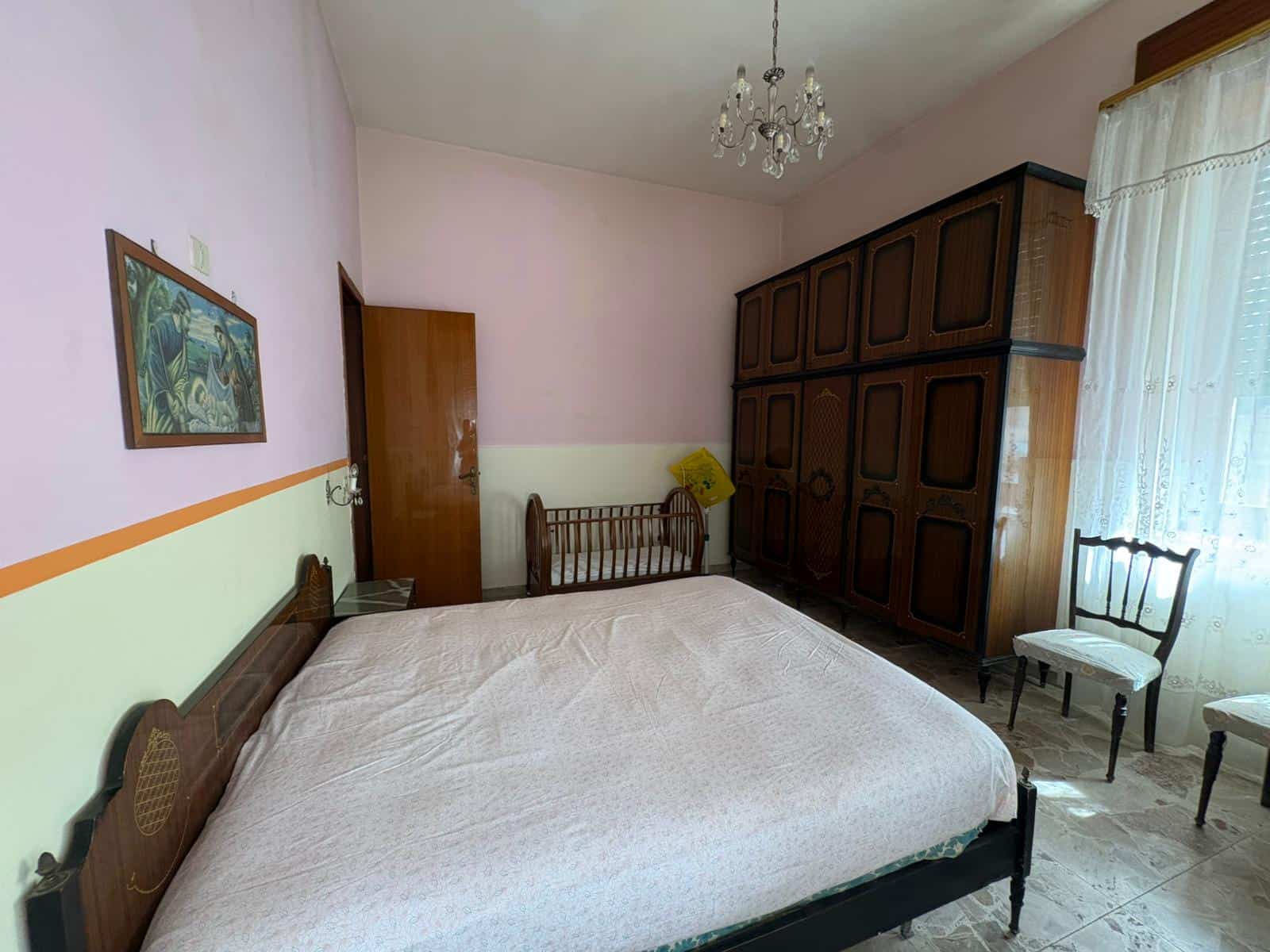Riproduzione di un'immagine di una camera da letto con arredi classici e pareti dipinte in tonalità pastello, illuminata da luce naturale attraverso una finestra con tende leggere.