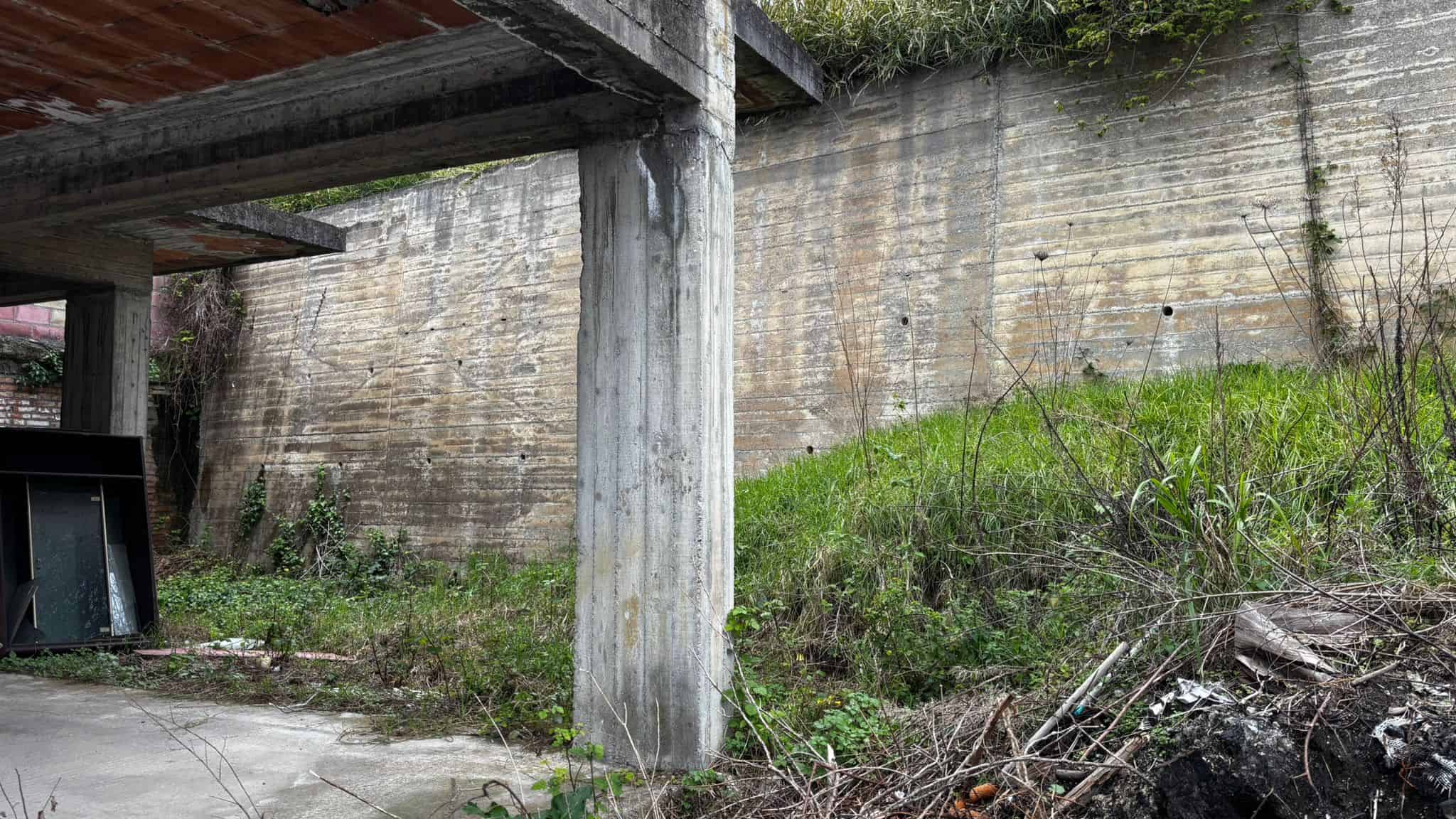 Scorcio di un'area esterna abbandonata con pareti in cemento e vegetazione spontanea, pronta per interventi di ristrutturazione o riqualificazione immobiliare.