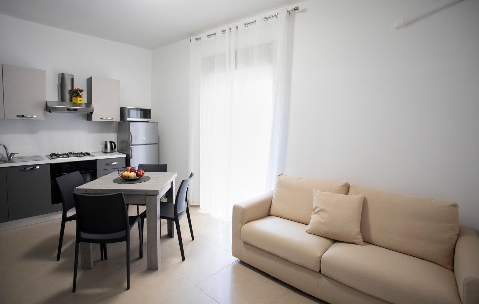 Interno di un soggiorno e cucina moderna con mobili grigi, divano beige e grande finestra con tende bianche, ambiente luminoso e accogliente.