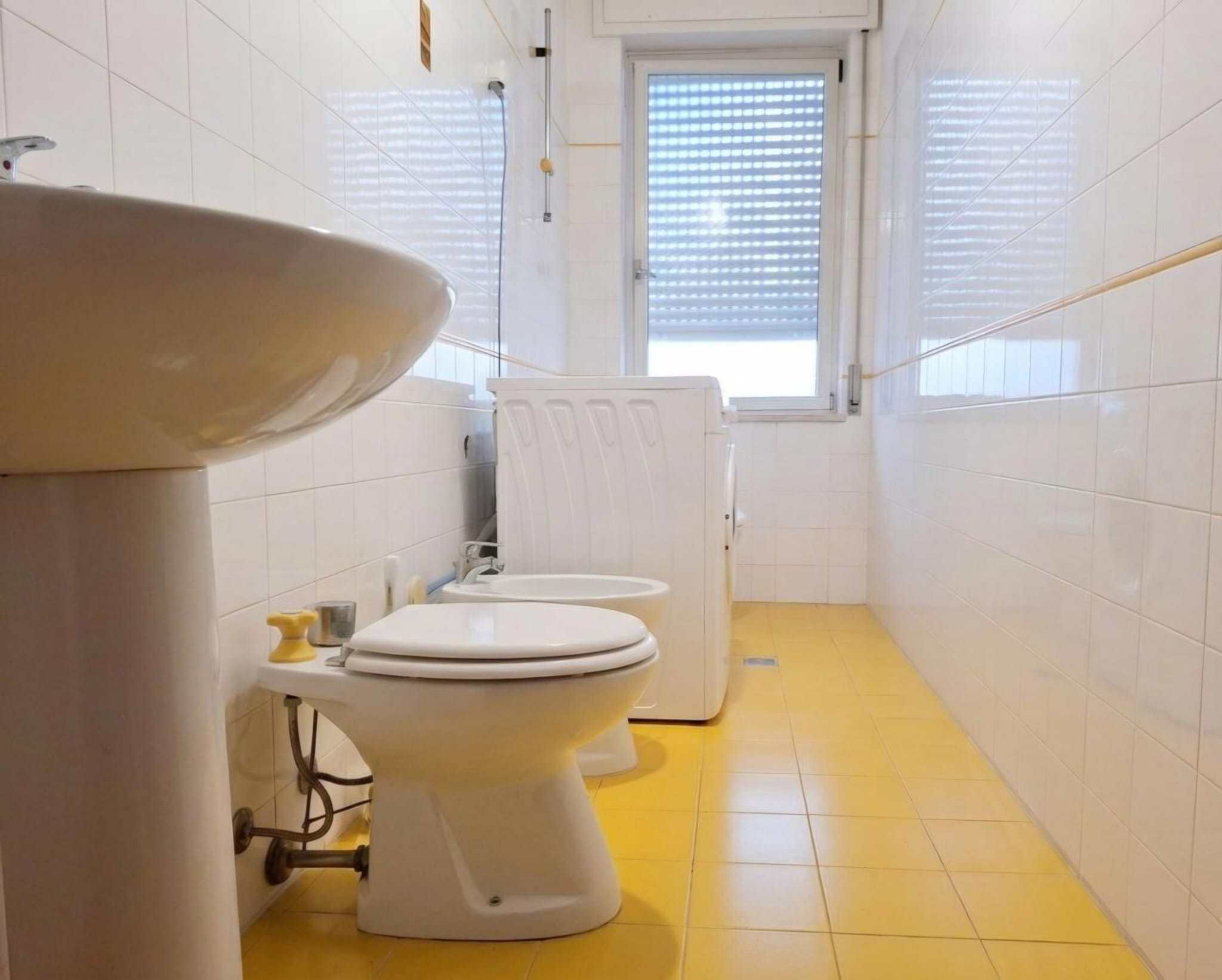 Lavabo e toilette in un bagno con pareti rivestite di piastrelle bianche e pavimento giallo, con finestra che permette l'ingresso di luce naturale. Ideale per ambienti di servizio o bagni di piccole d.