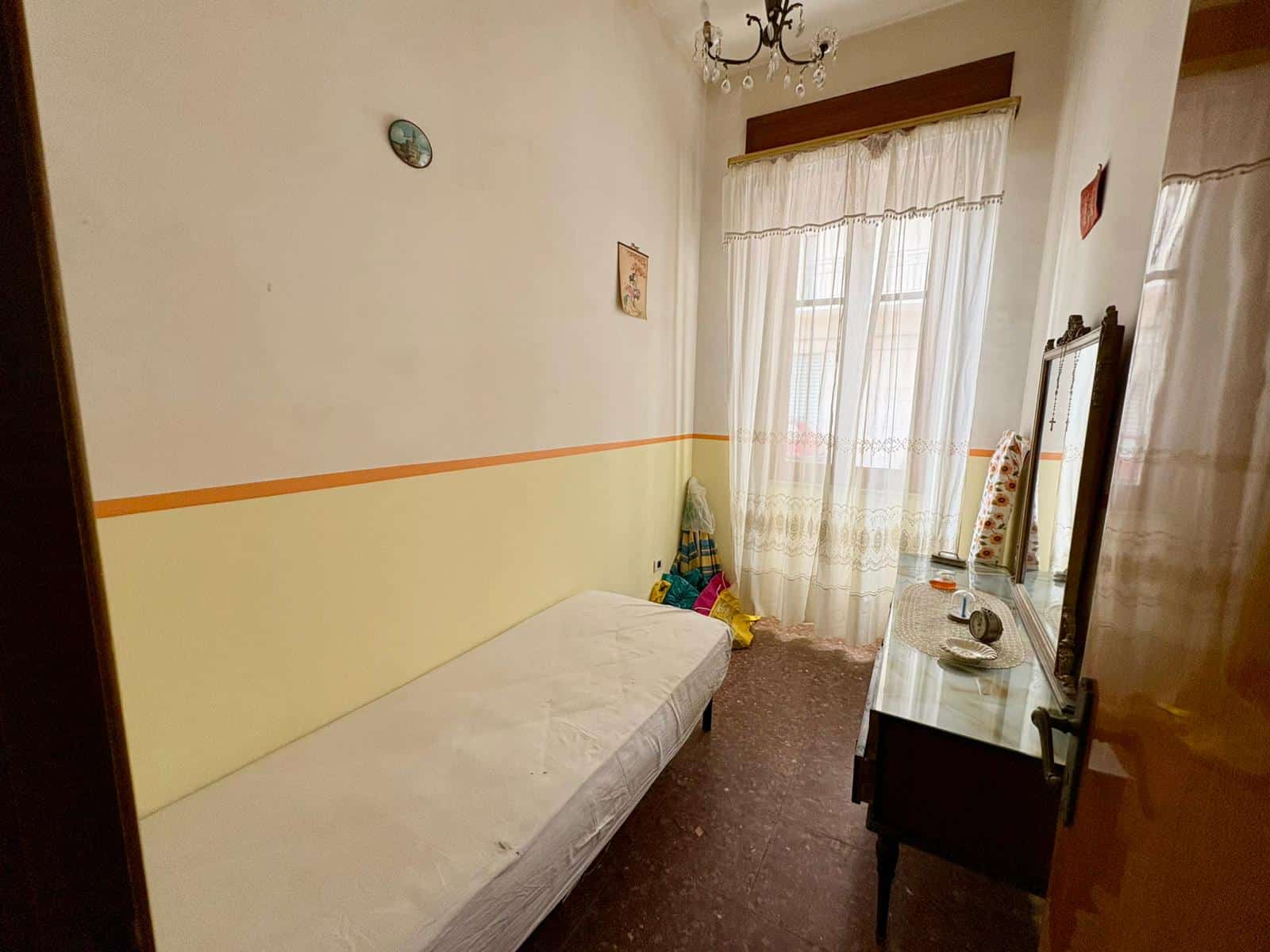 Letto singolo in camera con finestra luminosa, arredamento semplice e pareti chiare, ideale per affitto o uso temporaneo.