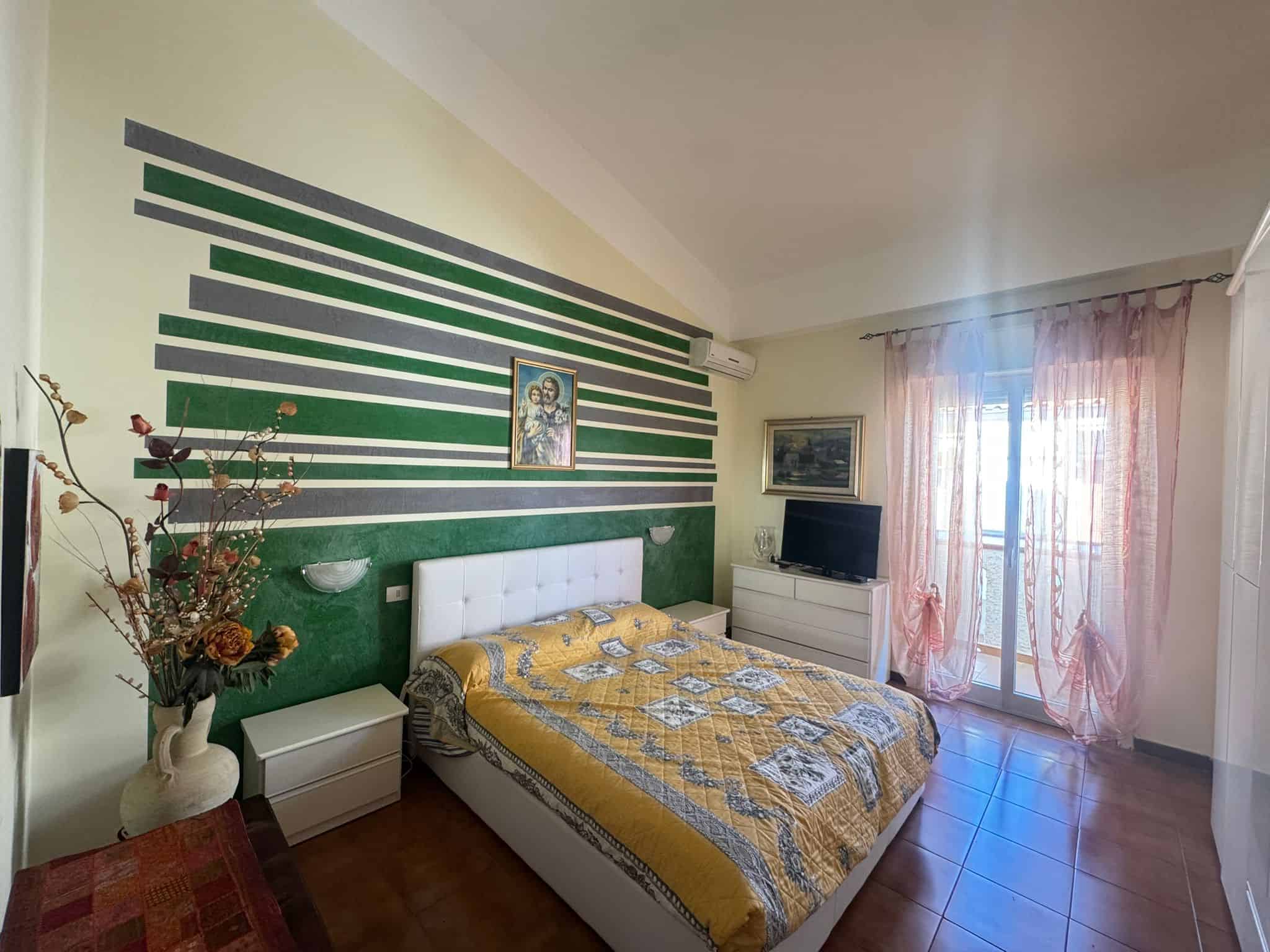 Ampia camera da letto moderna con pareti decorate con strisce verdi e bianche, letto matrimoniale con biancheria gialla, finestra con tende rosa e balcone, arredi eleganti e atmosfera accogliente.