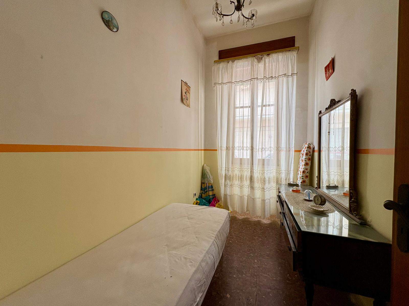 Una stanza singola luminosa con pareti bianche e gialle, finestra con tende leggere, letto singolo e specchio grande su una scrivania in legno, ambiente semplice e accogliente.