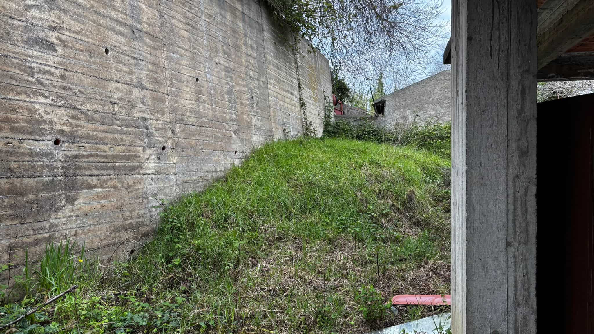 Verde pendio con erba e piante, circondato da muri in cemento e strutture in legno, in un'area esterna di un edificio residenziale o commerciale.