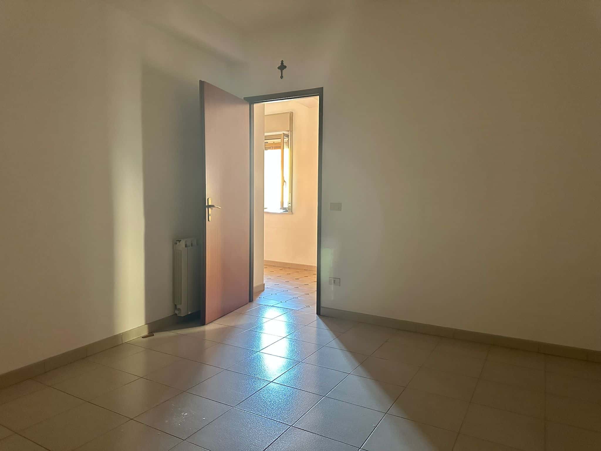 Una stanza vuota con pareti bianche, pavimento in piastrelle e porta aperta che conduce a una stanza luminosa con finestra. Ideale per annunci immobiliari e ristrutturazioni.