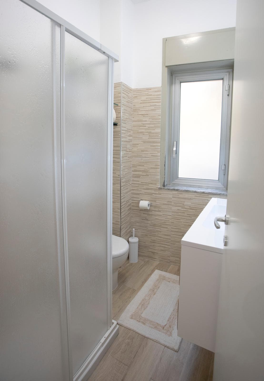 Pannello doccia in vetro opaco, bagno compatto con finestra, lavabo con mobiletto e accessori essenziali, arredo moderno e funzionale, ideale per spazi ridotti, design minimalista, ambiente luminoso e.