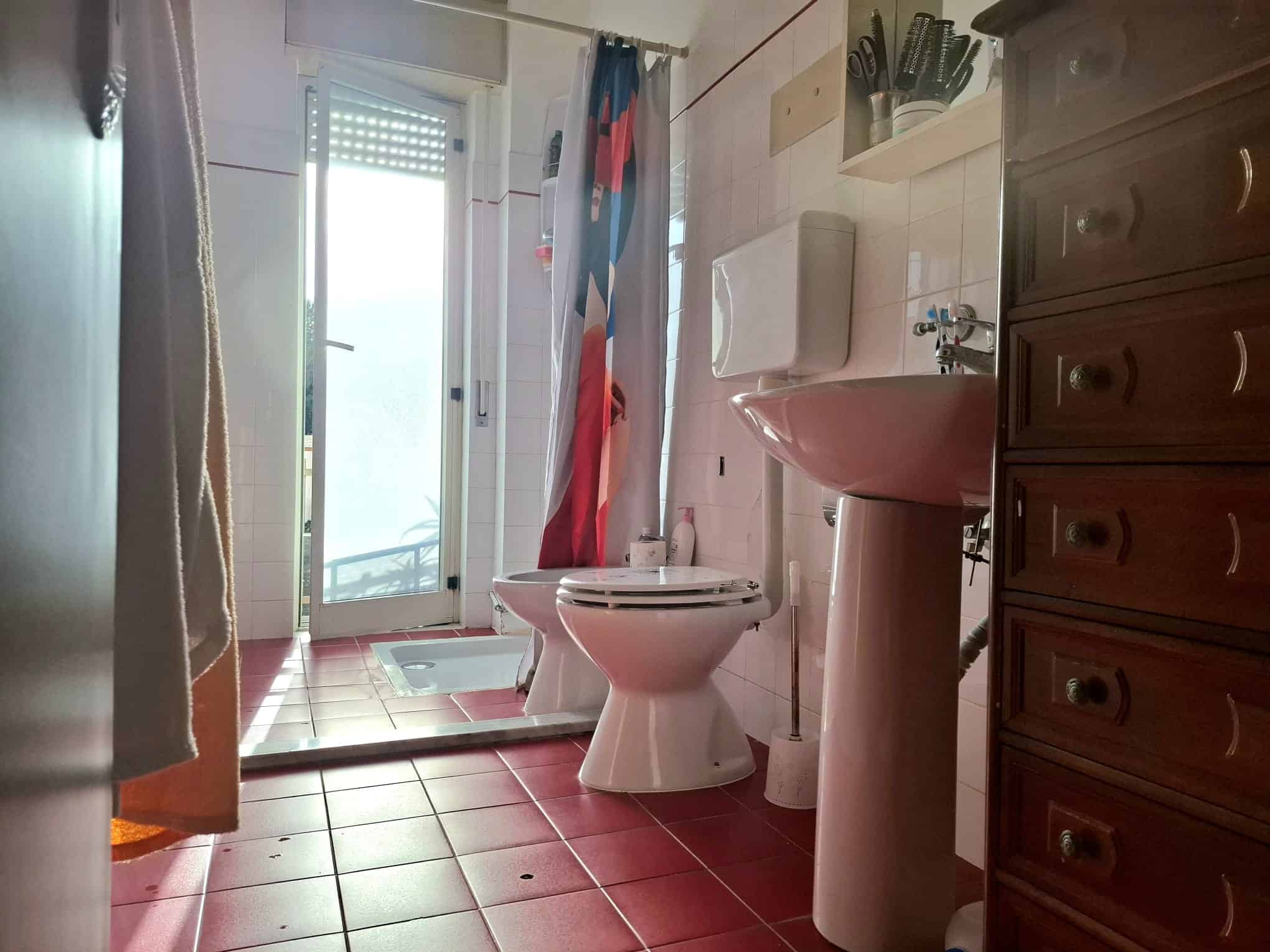 Lavabo e toilette in un bagno luminoso con accesso a un balcone, pavimento in piastrelle rosse e arredi in legno scuro, ideale per ambienti di case moderne e funzionali.