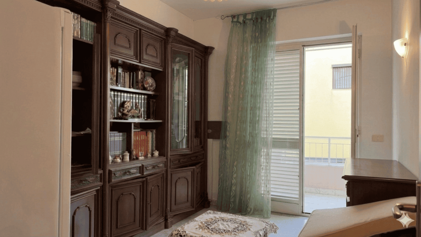 Ampio soggiorno con libreria in legno scuro, porta finestra con tende verdi e balcone, arredamento classico, ambiente luminoso e accogliente, ideale per vivere momenti di relax e convivialità.