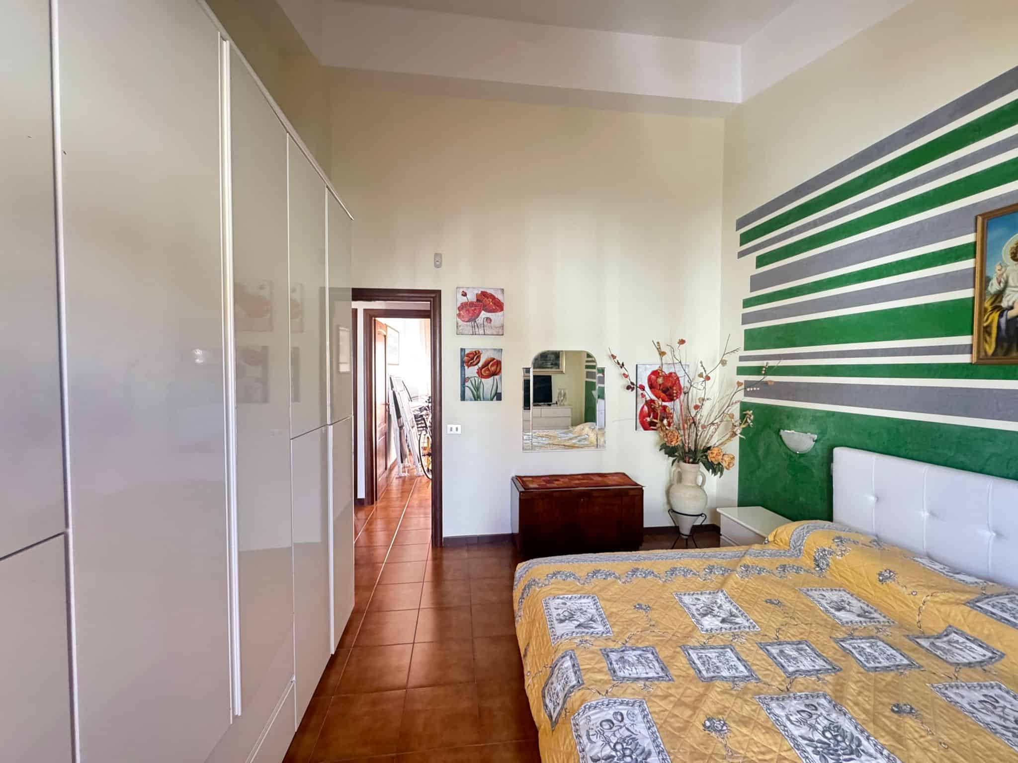 Letto matrimoniale con testiera imbottita e coperta colorata, in una camera da letto moderna con pareti decorate a righe verdi e bianche, armadio a muro e accessori decorativi.