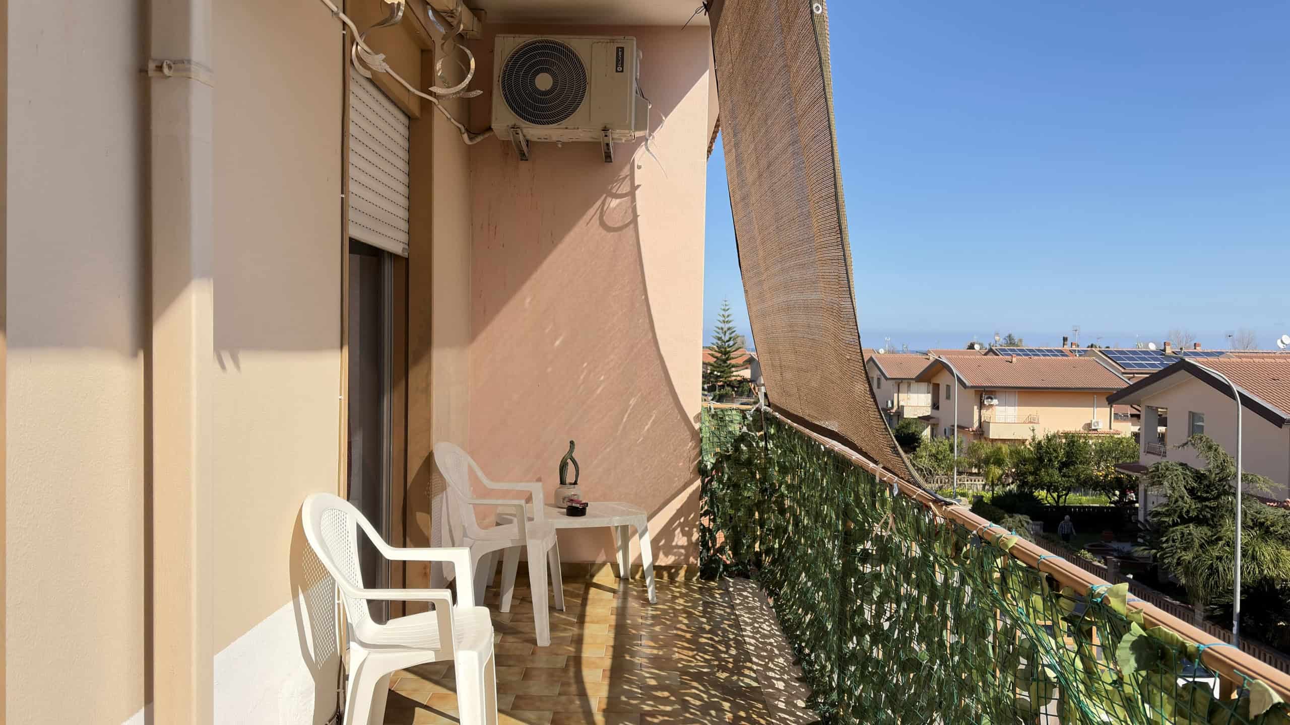 Balcone di un appartamento con vista su case e cielo sereno, arredato con sedie e tavolino, decorato con piante verdi, ideale per relax e momenti all'aperto.
