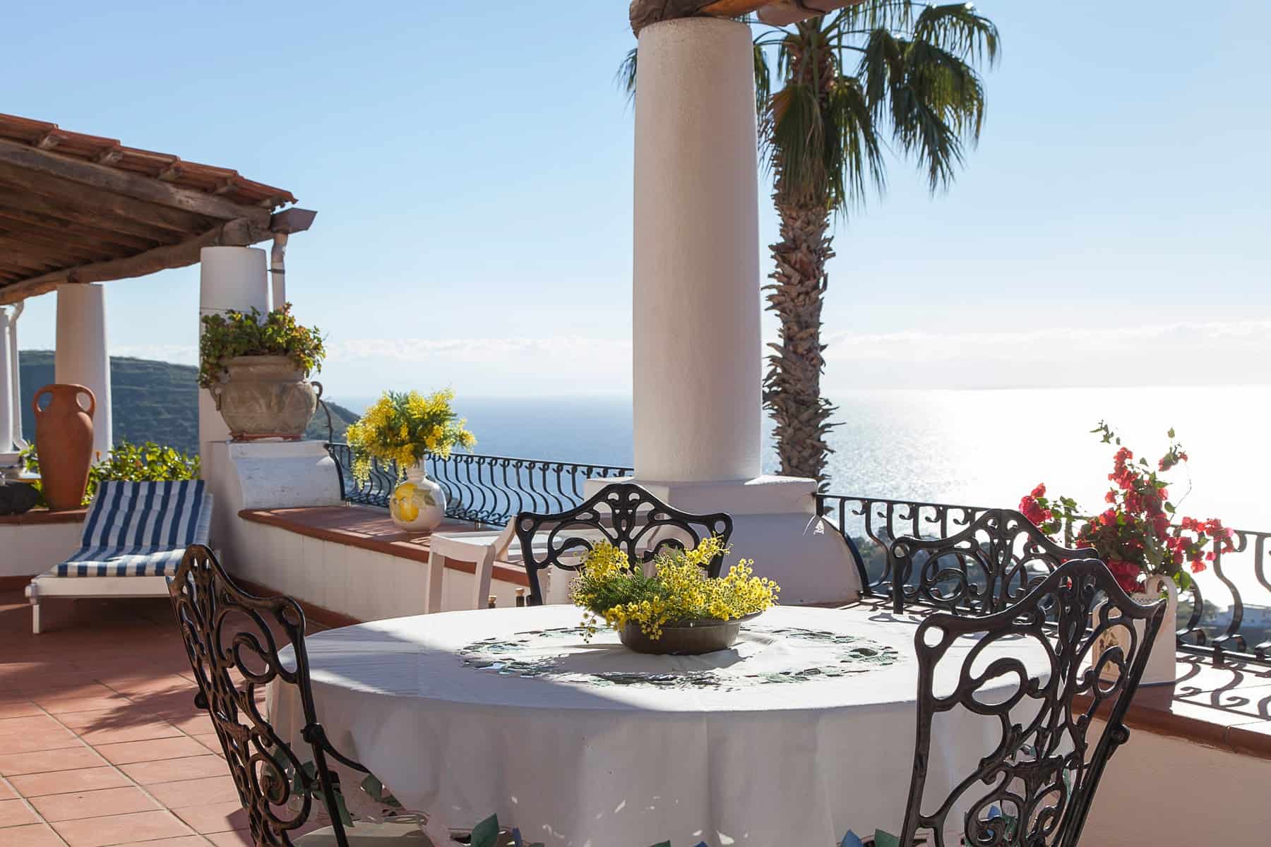 Piatto da pranzo all'aperto con vista panoramica sul mare, arredato con tavolo rotondo, sedie in ferro battuto e decorazioni floreali, ideale per relax e cene estive.