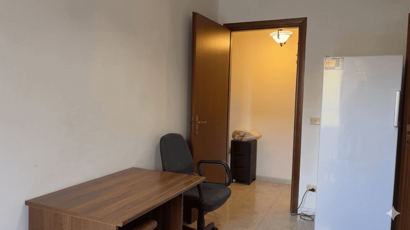 Porte aperte verso un ingresso di casa con arredamento semplice, vista da un soggiorno o camera, con mobili essenziali e ambiente luminoso.