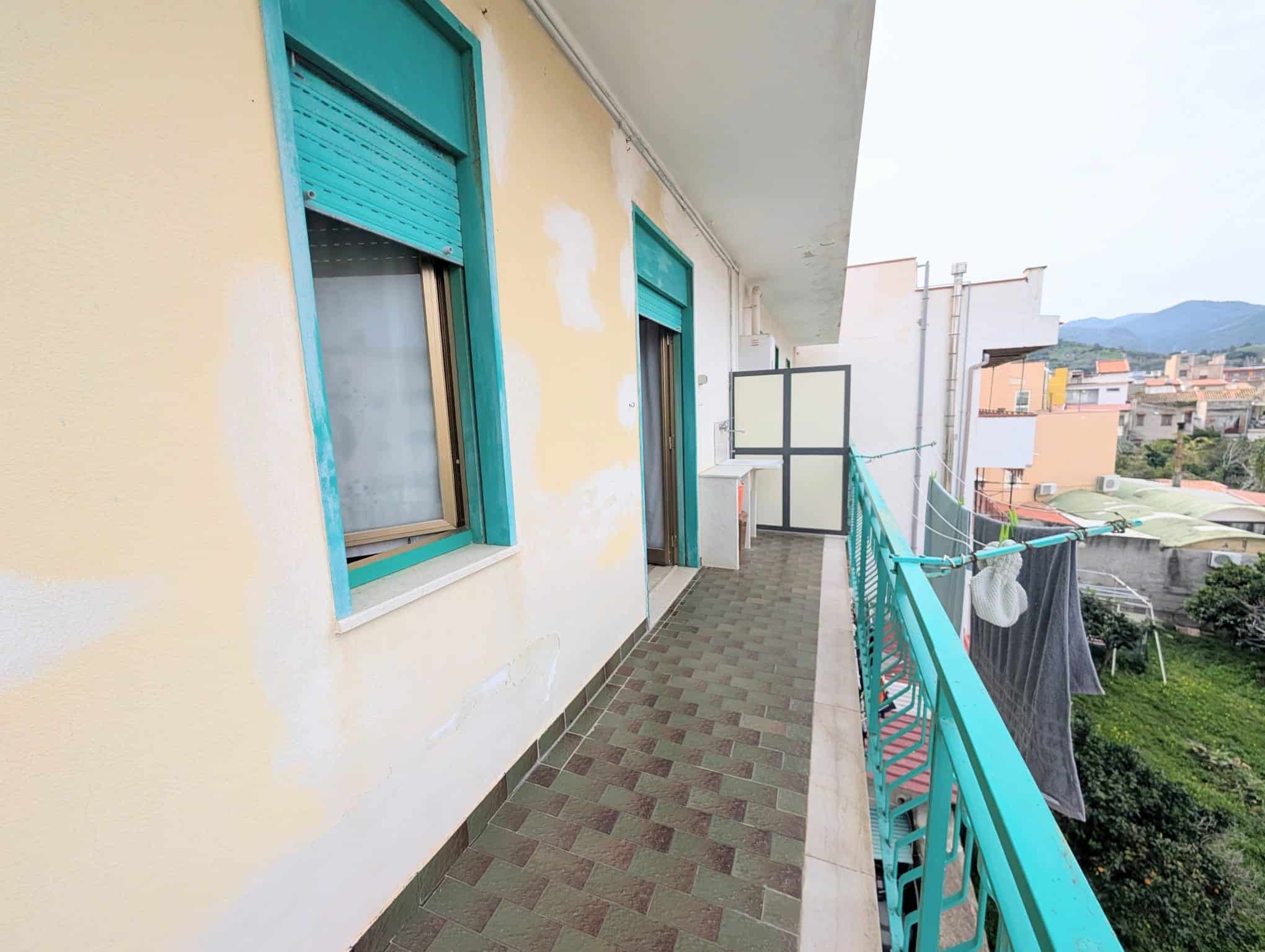 Aggiungi un balcone di un appartamento con vista sulla città e sulle colline circostanti, con pareti gialle e finestre con tapparelle azzurre.