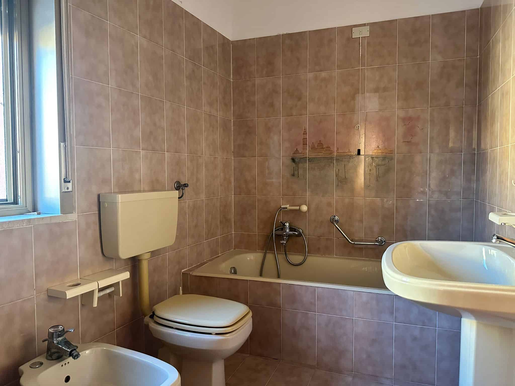Lavabo, WC e vasca da bagno in un bagno di casa con pareti rivestite di piastrelle beige, finestra per l'illuminazione naturale e accessori essenziali per l'igiene quotidiana.