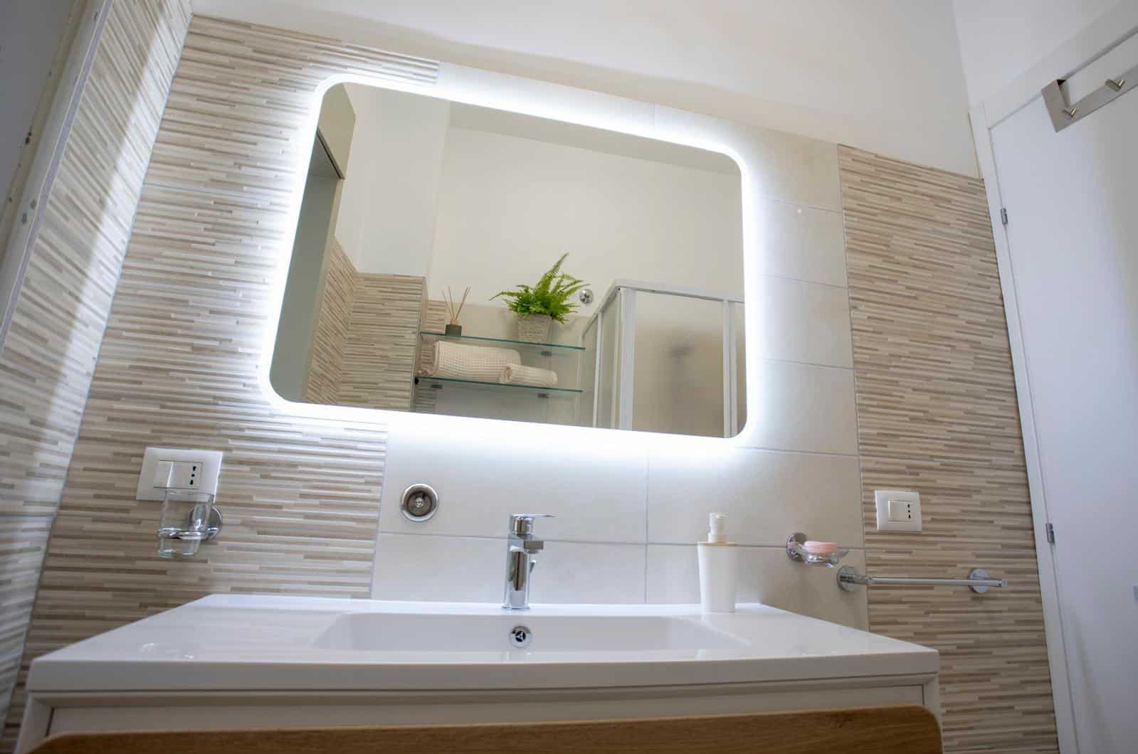 Lavabo moderno con grande specchio illuminato, pareti in piastrelle beige e dettagli minimalisti, ideale per bagni di design e funzionali.