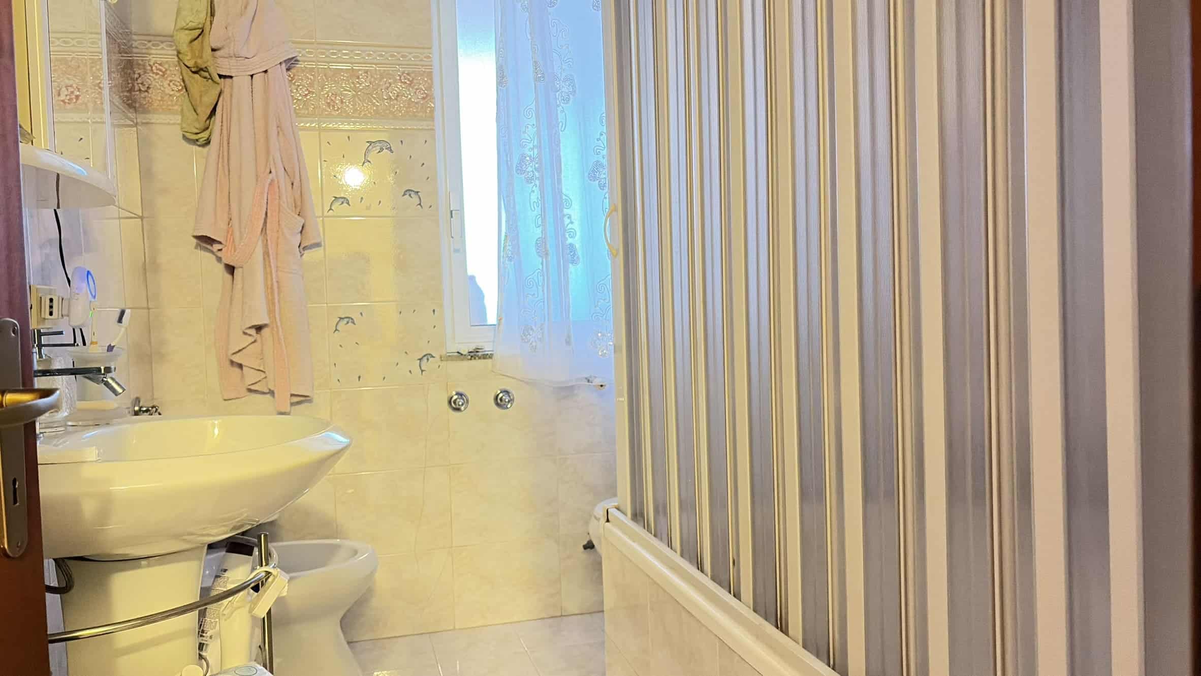 Spazio bagno moderno con doccia in vetro, lavabo e accessori, illuminato da luce naturale attraverso una finestra con tende leggere, ambiente pulito e funzionale.