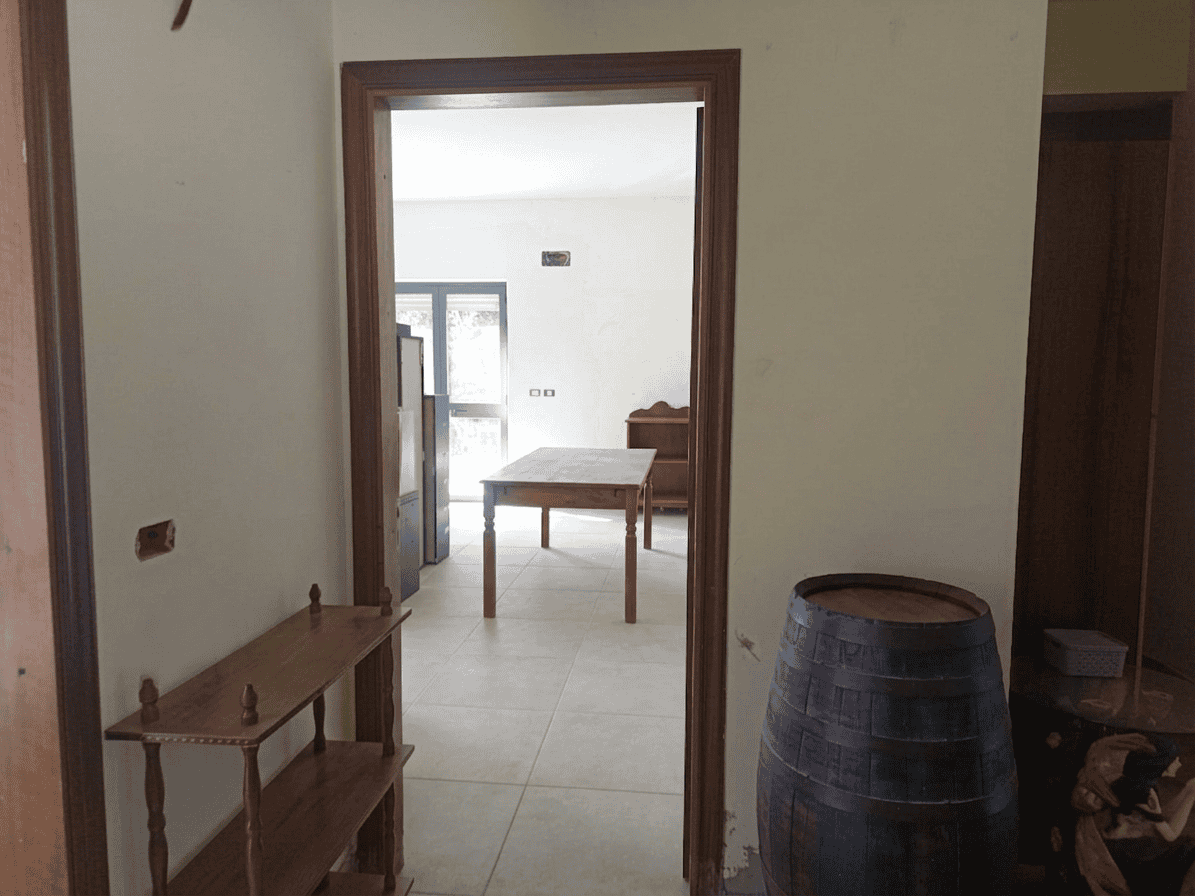 Vista di un ingresso in un appartamento con pareti bianche, pavimento in piastrelle chiare e mobili in legno, con un grande barile decorativo in primo piano a destra.