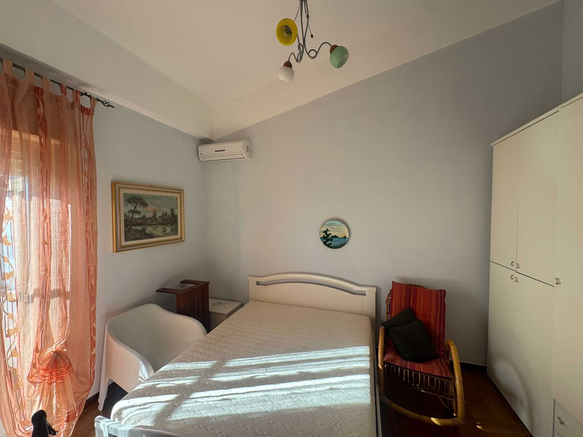 Letto matrimoniale in una camera luminosa con pareti azzurro chiaro, finestra con tende di pizzo rosa, arredi moderni e decorazioni marine, ideale per affitti turistici o residenziali.