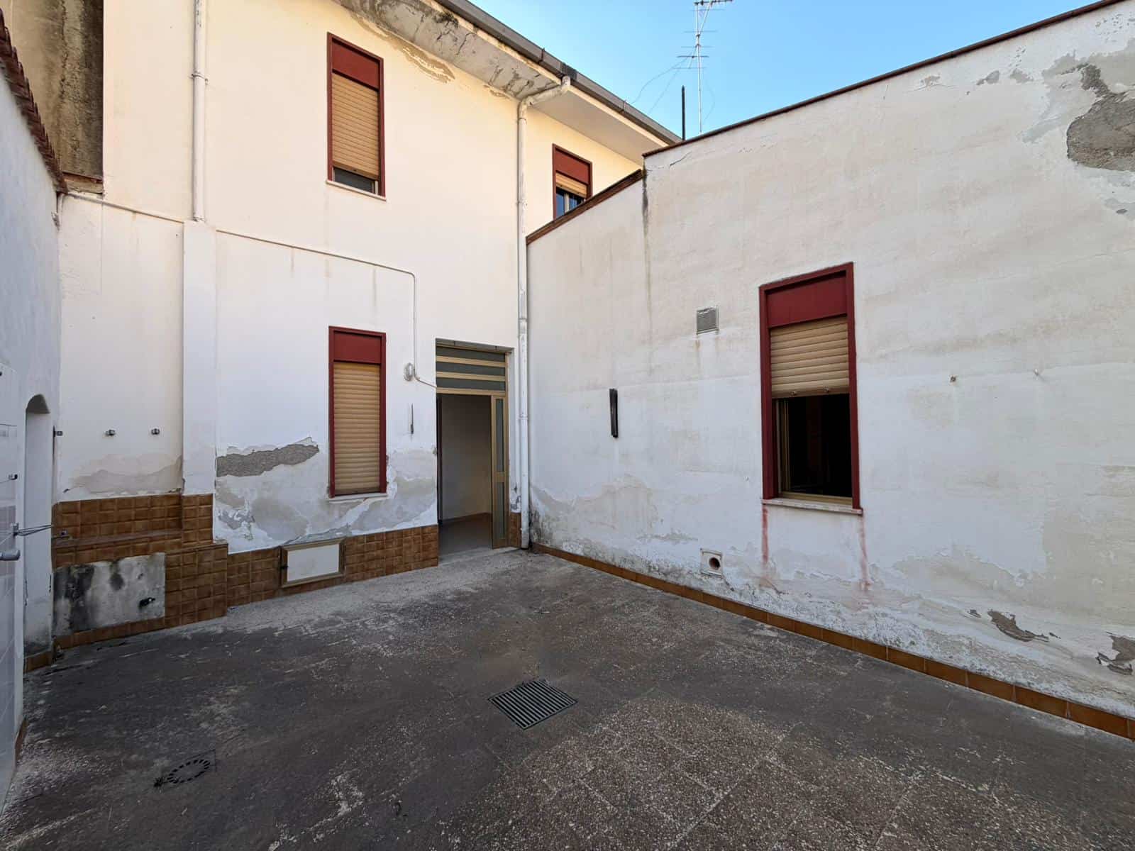 Cortile interno di un edificio residenziale con pareti bianche e finestre con tapparelle rosse, area esterna pavimentata, ideale per ristrutturazione o utilizzo come spazio esterno privato.