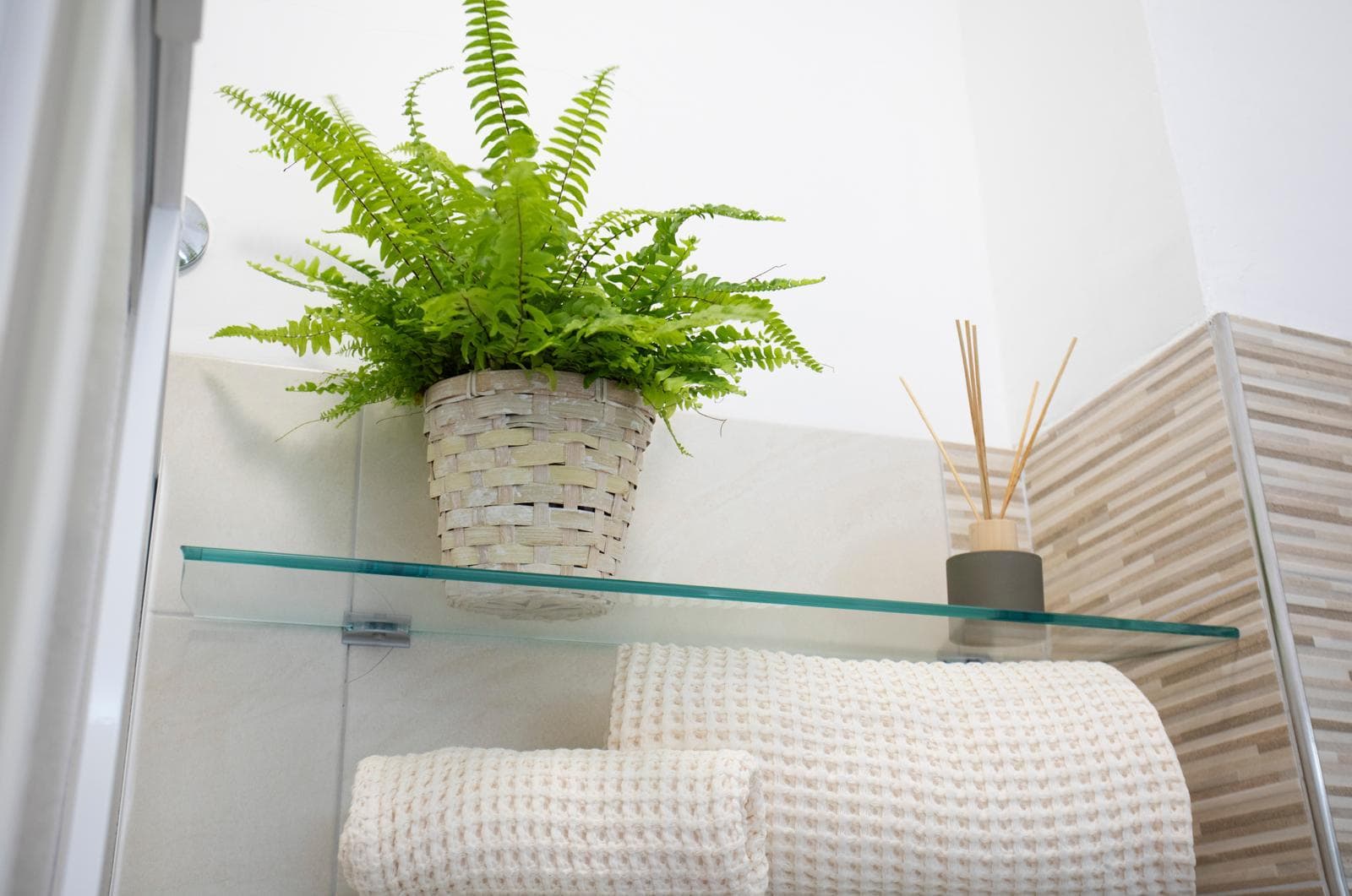 Piante verdi in vaso decorativo su mensola di vetro in bagno, con asciugamani bianchi e diffusore di fragranze, ambiente luminoso e moderno.