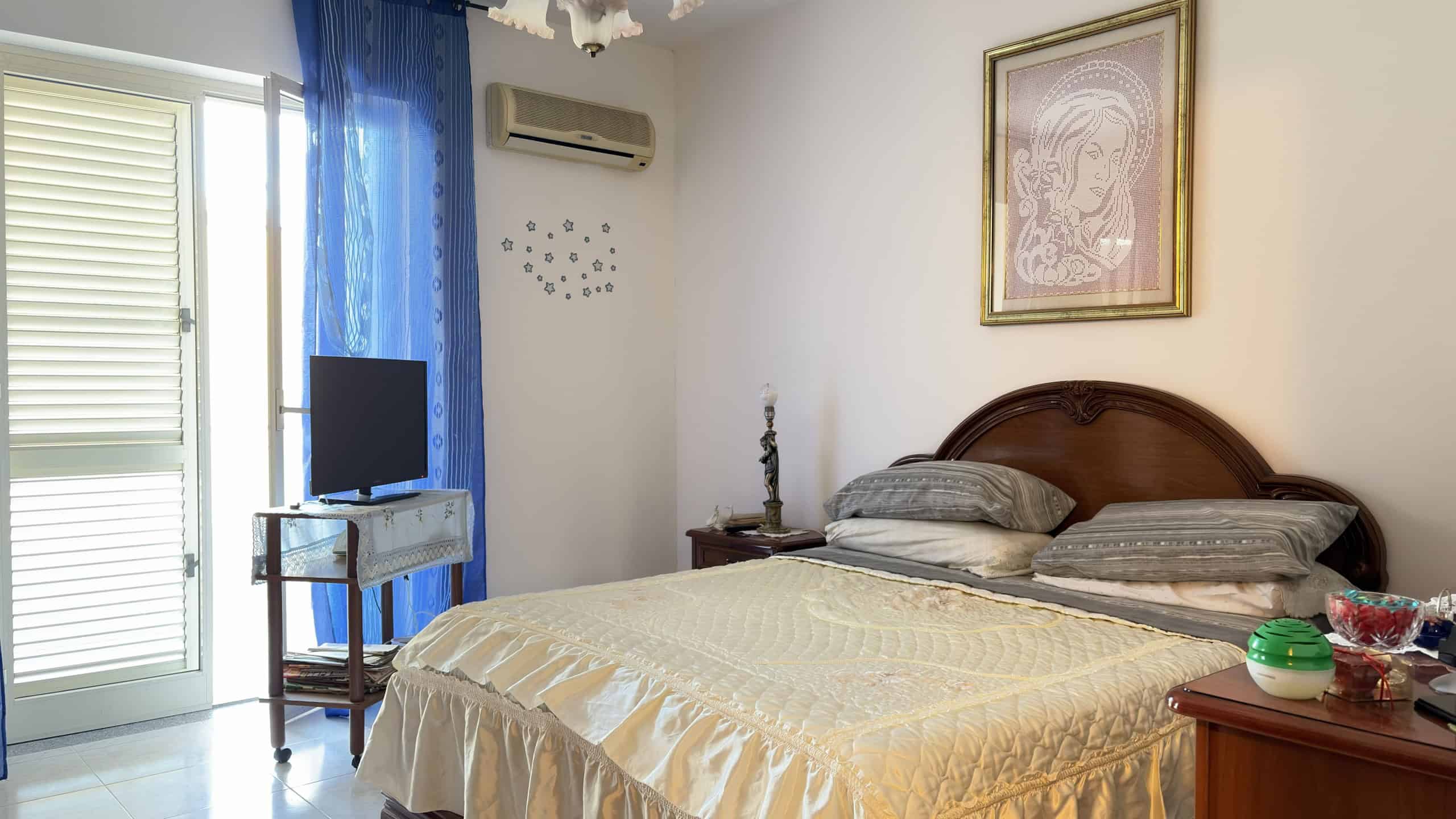 Letto matrimoniale con testiera in legno, in una camera da letto luminosa e accogliente, decorata con quadri e tende blu, ideale per ambienti di comfort e relax.