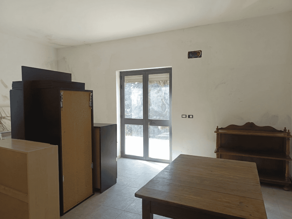 Interno di una stanza vuota con porta finestra, mobili di legno e pareti bianche, pronta per ristrutturazione o arredamento.