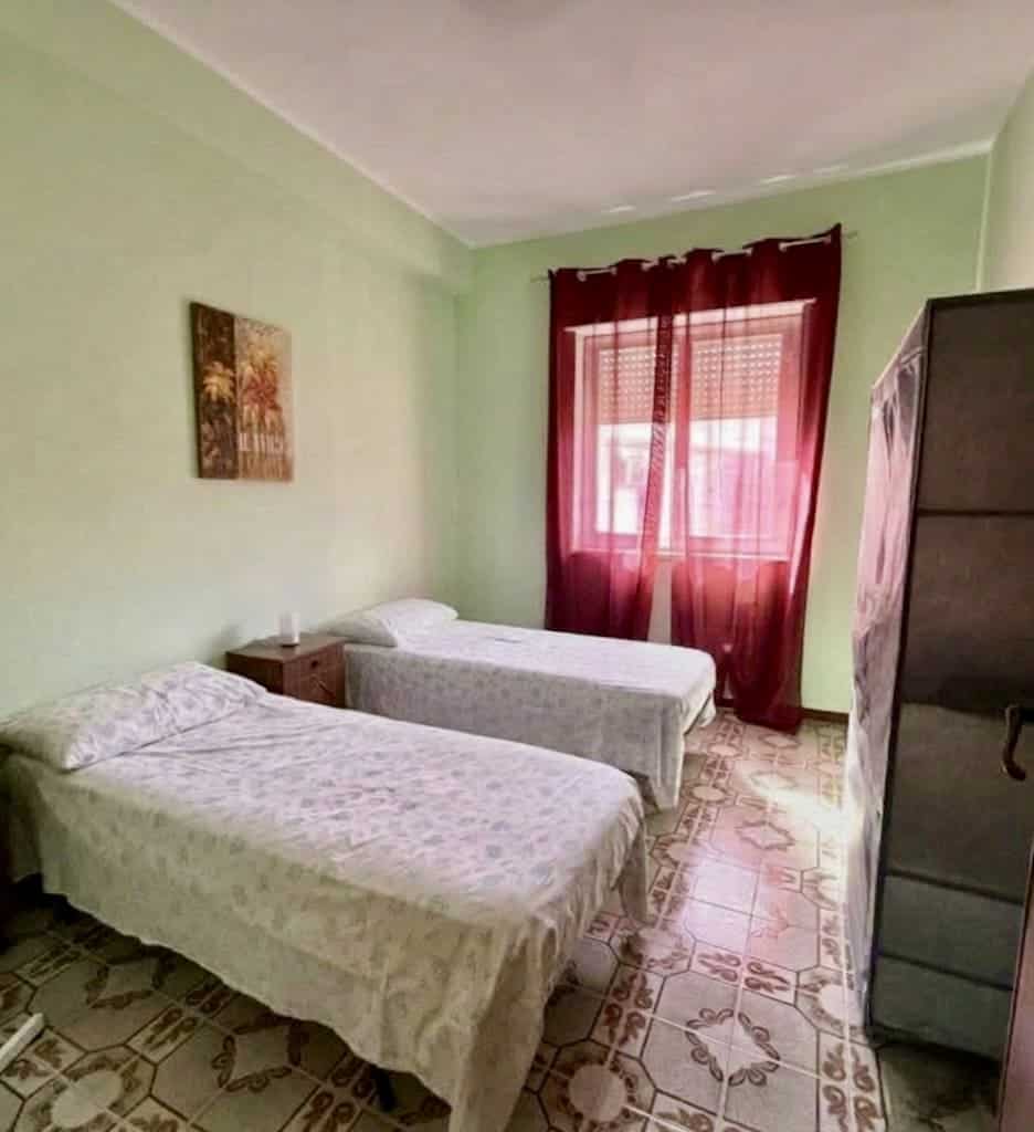 Camera da letto con due letti singoli, pareti verde chiaro, finestra con tende rosse, arredi semplici, ideale per affitti brevi o soggiorni familiari.