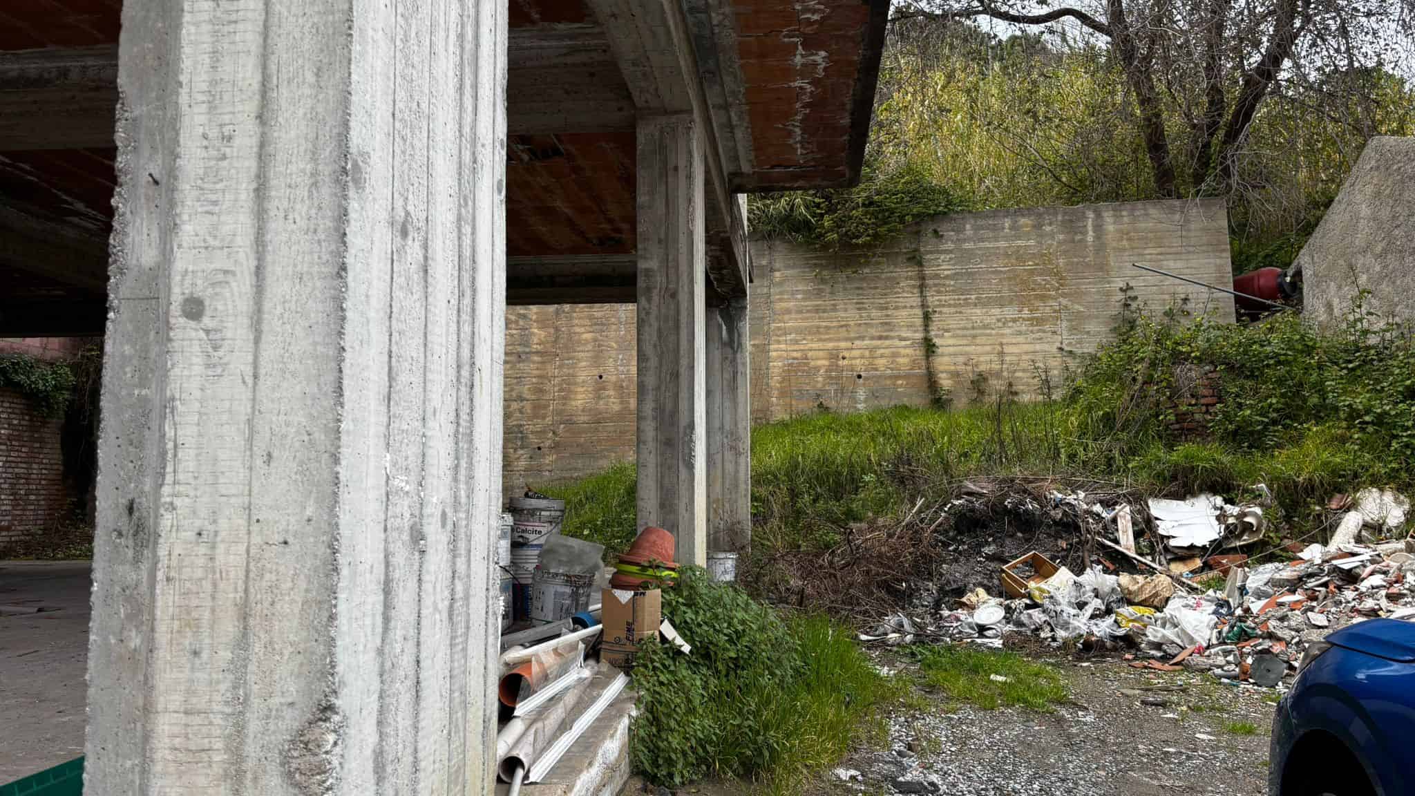 Scorcio di un'area esterna abbandonata con rifiuti e detriti sotto un edificio in cemento, ambiente trascurato e degrado urbano.