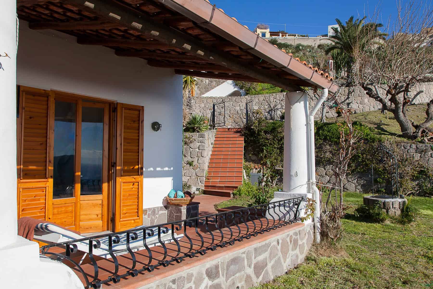 Vicolo di una casa con veranda in stile mediterraneo, con porte in legno e balcone in ferro battuto, circondata da un giardino con alberi e piante, in un contesto tranquillo e soleggiato.
