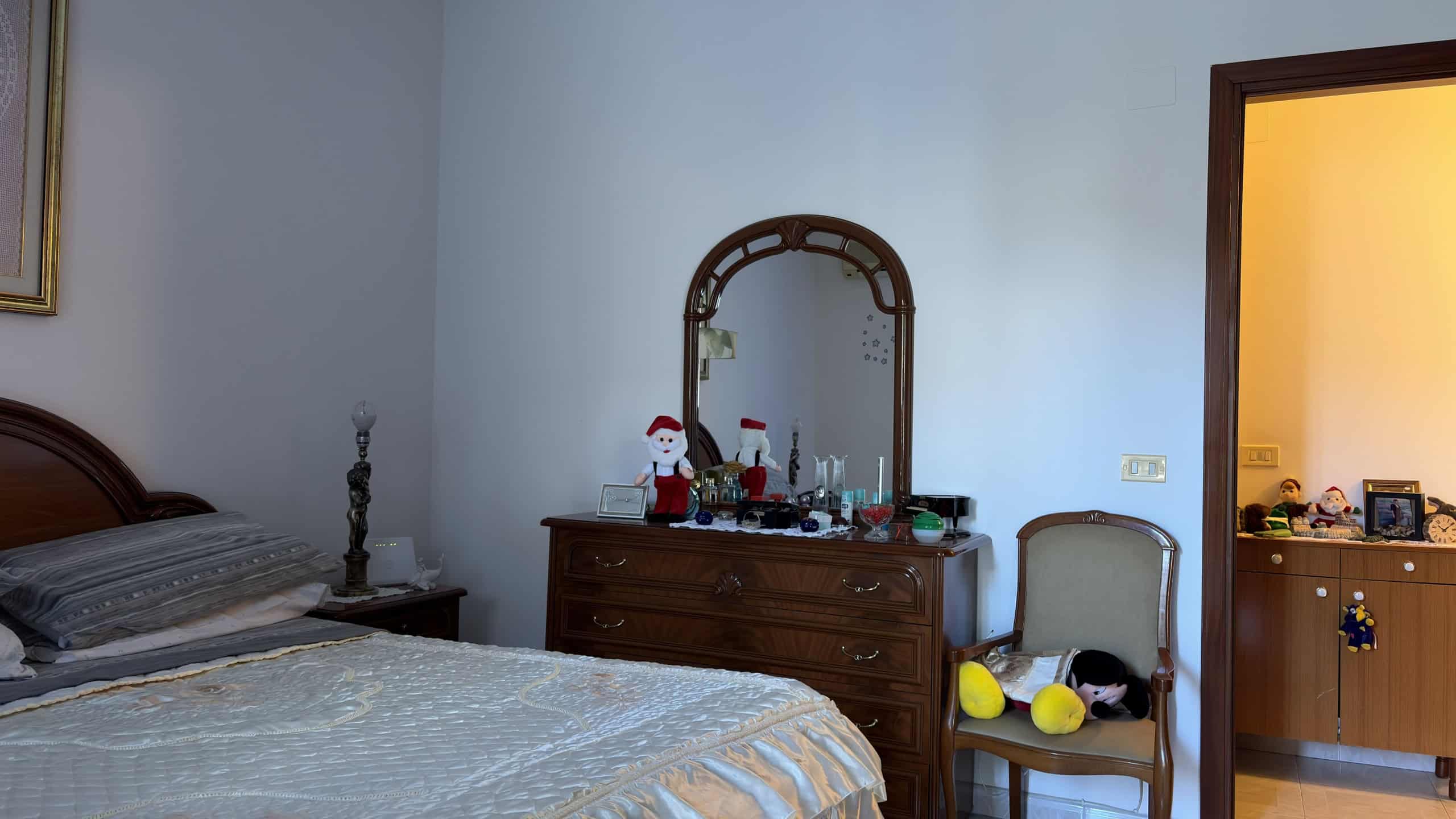 Ampia camera da letto con letto matrimoniale, comò in legno con specchio, decorazioni natalizie e ambiente accogliente. Perfetta per case in vendita o affitto, con stile classico e comfort.