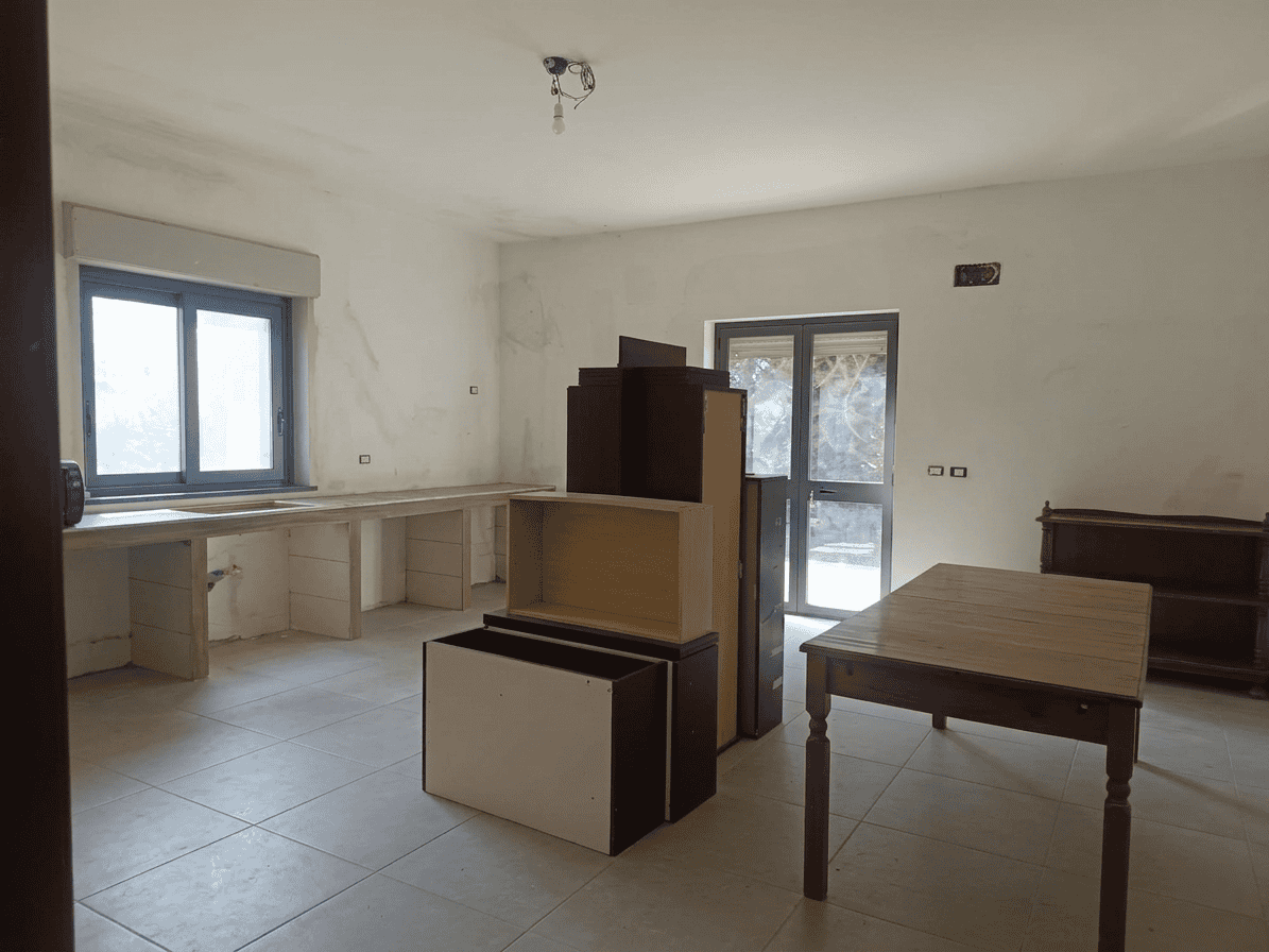 Il testo dell'immagine mostra un interno di una casa in fase di ristrutturazione, con pareti non verniciate e mobili temporanei, illuminata da luce naturale proveniente dalle finestre.