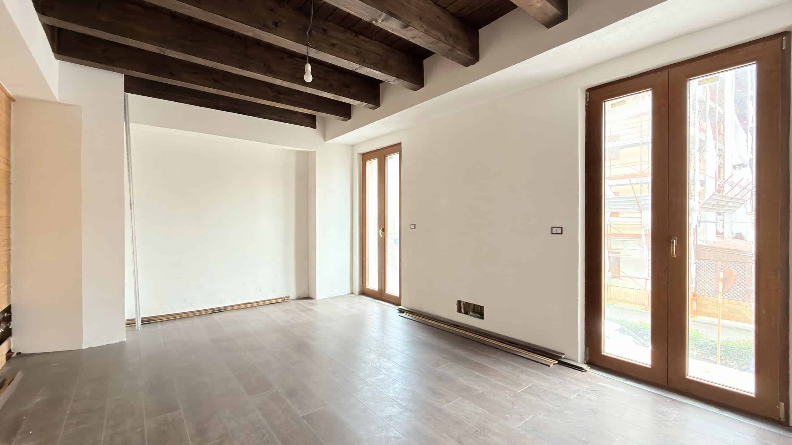 Ampia stanza luminosa con pareti bianche, soffitto con travi in legno a vista e grandi finestre in legno che lasciano entrare molta luce naturale, ideale per un soggiorno moderno e accogliente.