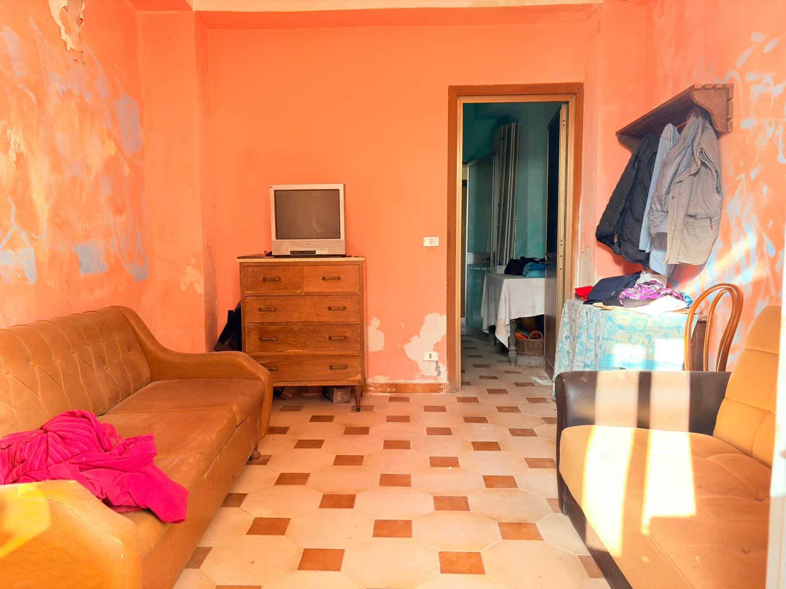 Soggiorno con pareti arancioni, divano in pelle marrone, armadio con TV, e ingresso a una stanza da letto, in un appartamento abitato.