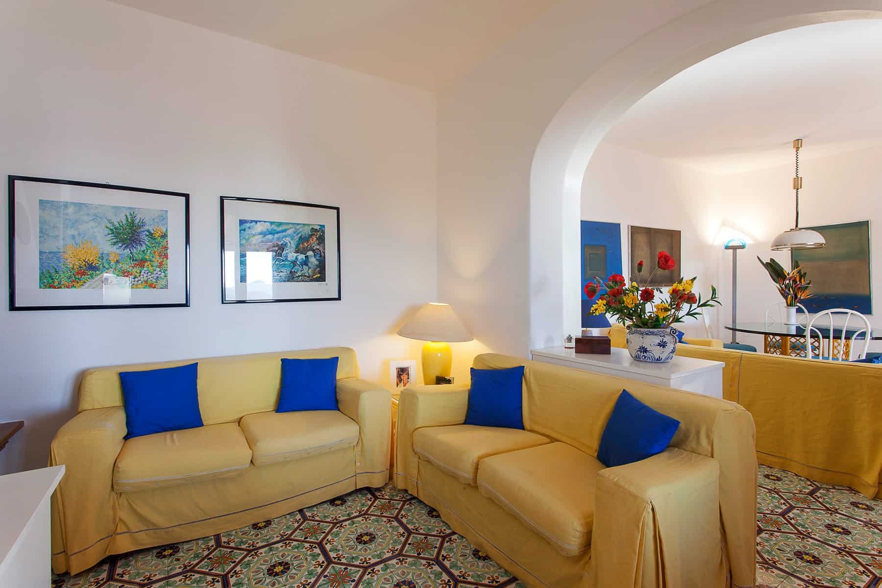 Un soggiorno luminoso con divani gialli e cuscini blu, decorato con quadri colorati e un tappeto elegante, ideale per vivere momenti di relax e convivialità in un ambiente accogliente.