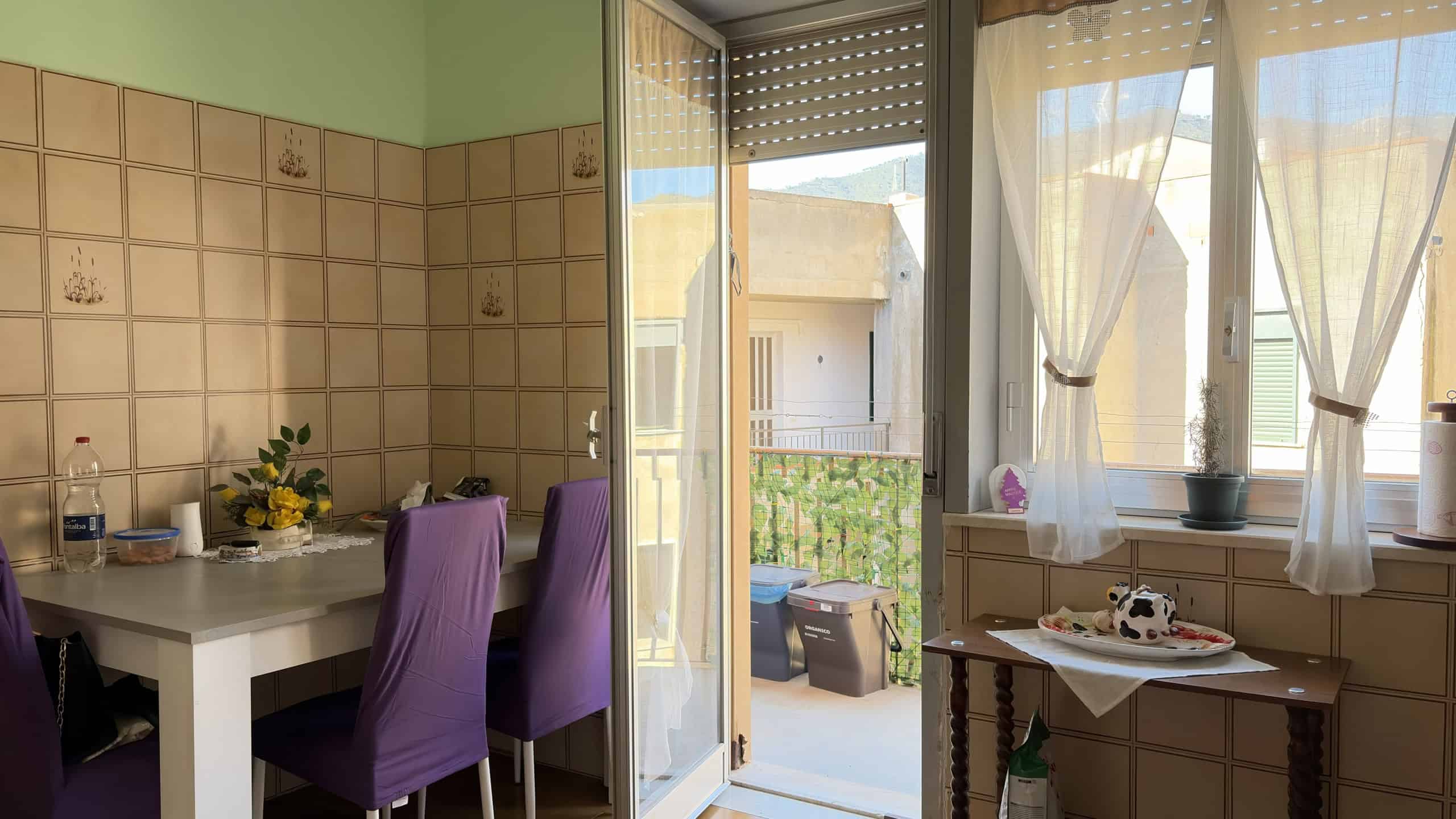 Vista di una cucina luminosa con balcone, pareti verdi e sedie viola, ideale per ambienti domestici accoglienti e moderni.