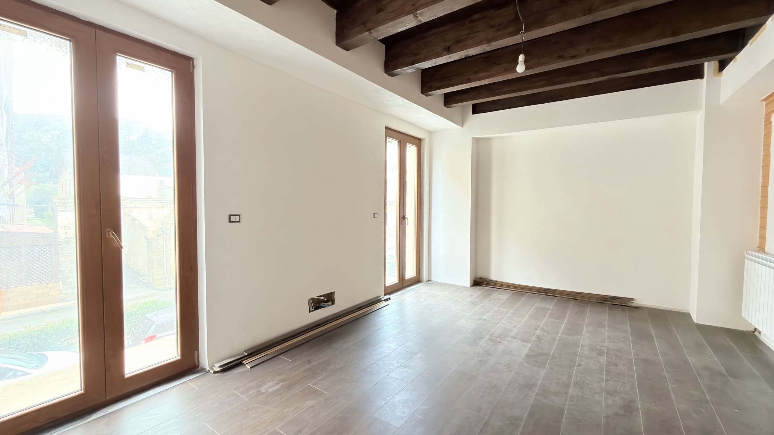 Ampia stanza luminosa con pareti bianche, soffitto con travi in legno a vista e grandi finestre che lasciano entrare molta luce naturale, ideale per arredamento moderno e accogliente.