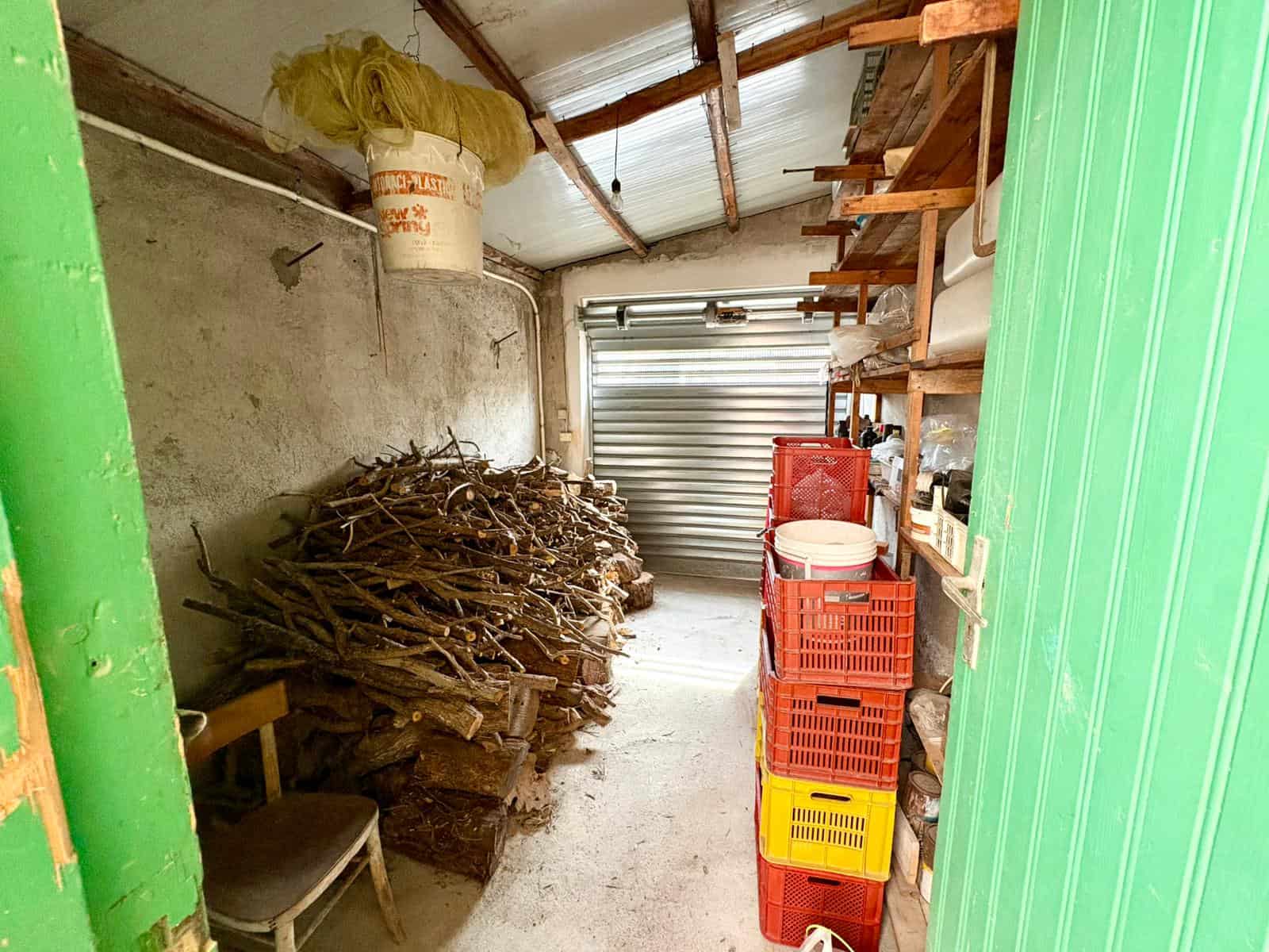 L'immagine mostra un garage con legna da ardere impilata, scaffalature con vari oggetti e un portone in metallo, rappresentando un ambiente di deposito e stoccaggio domestico.