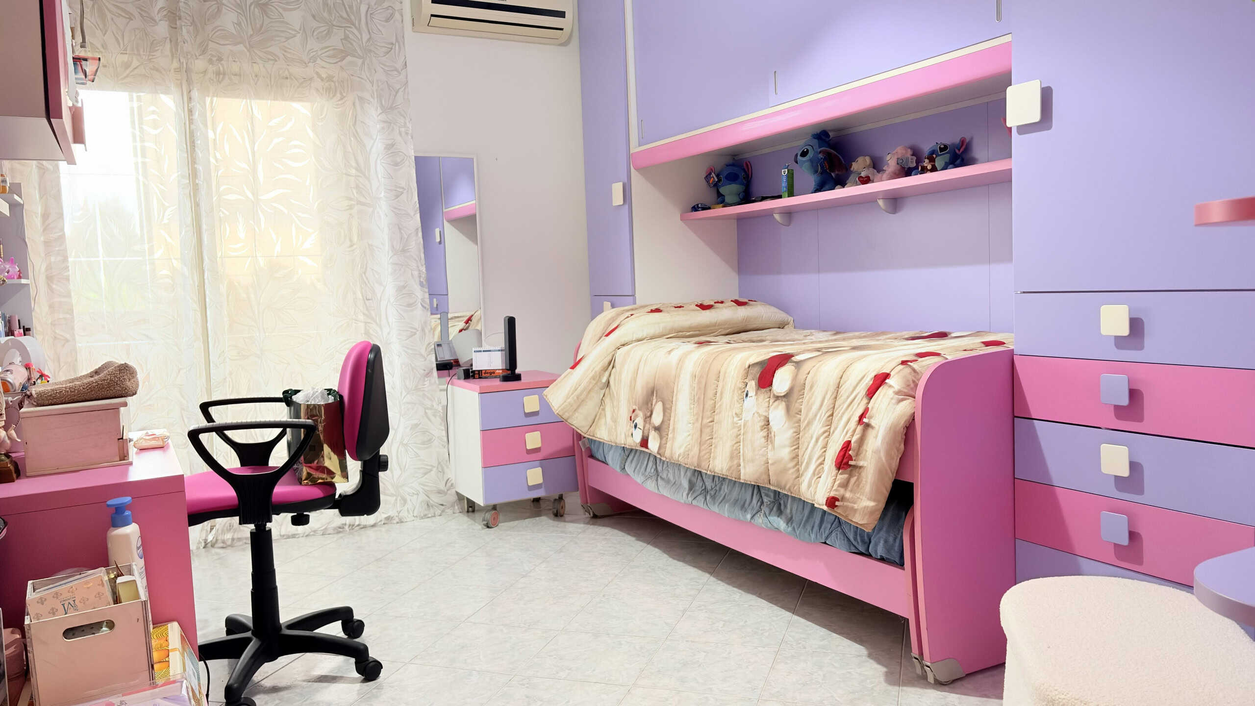 Lettino singolo colorato in una cameretta moderna con mobili rosa e lilla, decorazioni giocose e spazio per studio e svago, ideale per bambini o adolescenti.