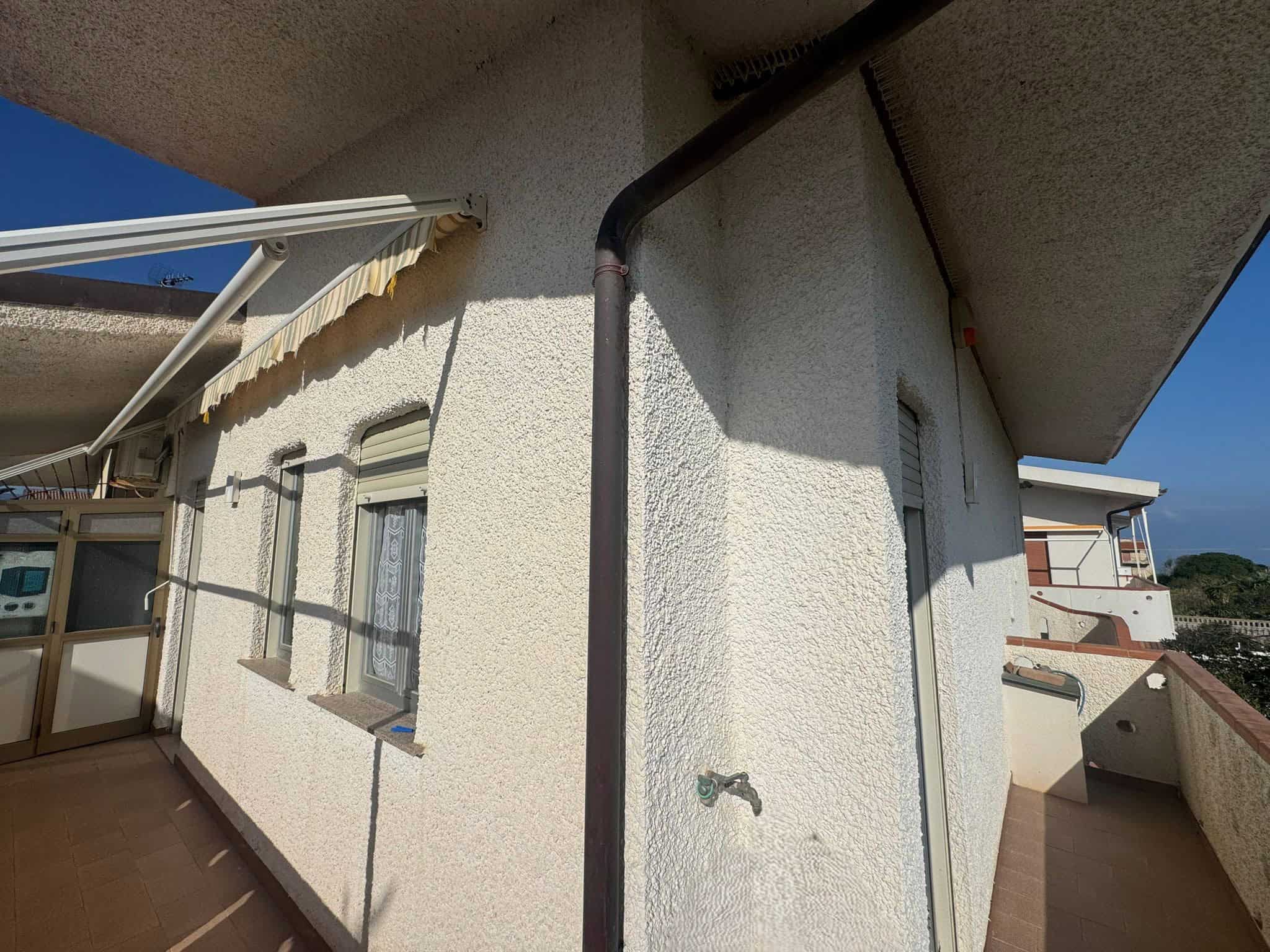 Balcone di un appartamento con pareti bianche, finestre e tende parasole, vista panoramica sulla città e il mare, ideale per case in affitto o residenze estive in Italia.